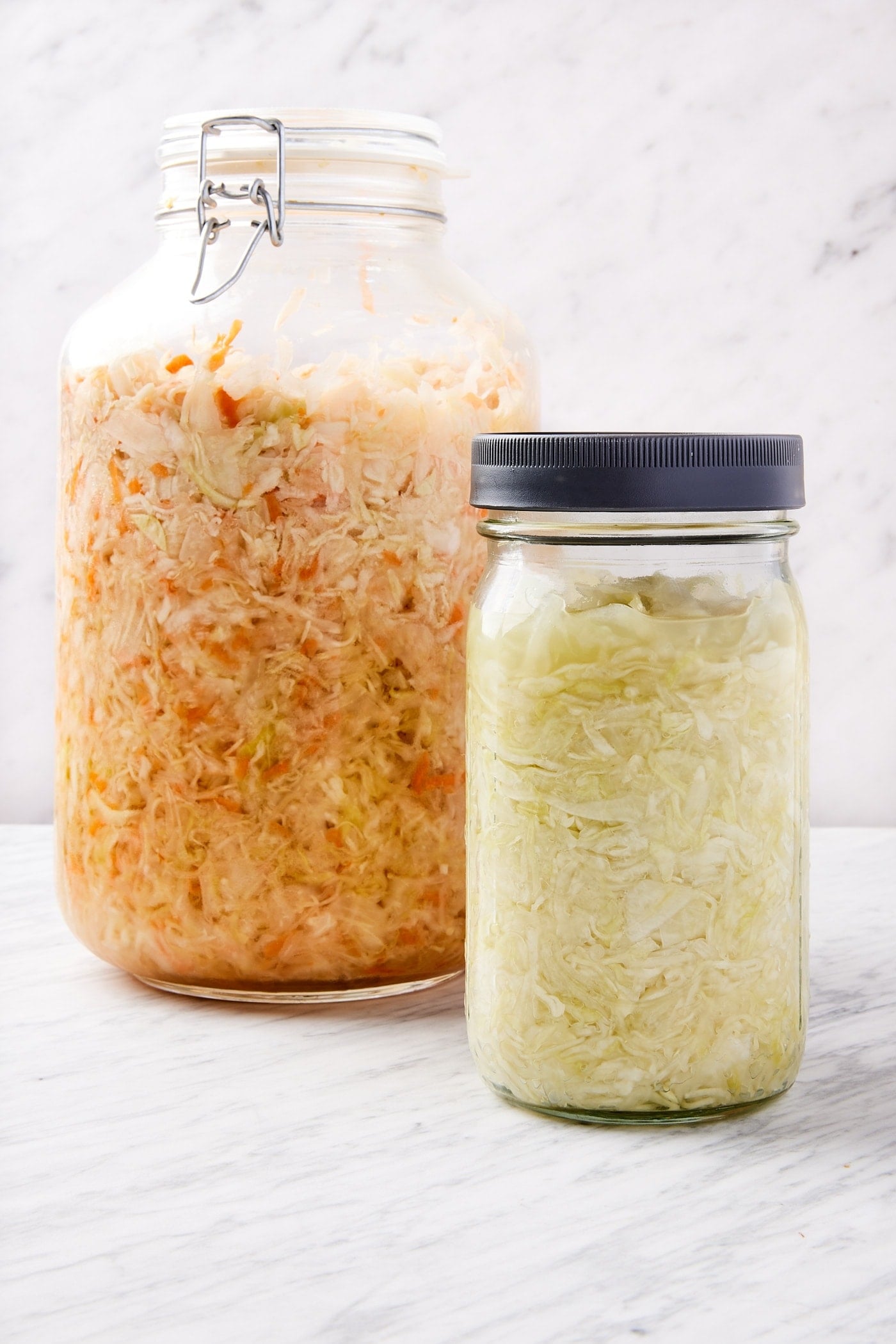 Two jars of homemade sauerkraut sitting on marble surface with white background with one jar filled with carrot along with sauerkraut and the other smaller jar plain.