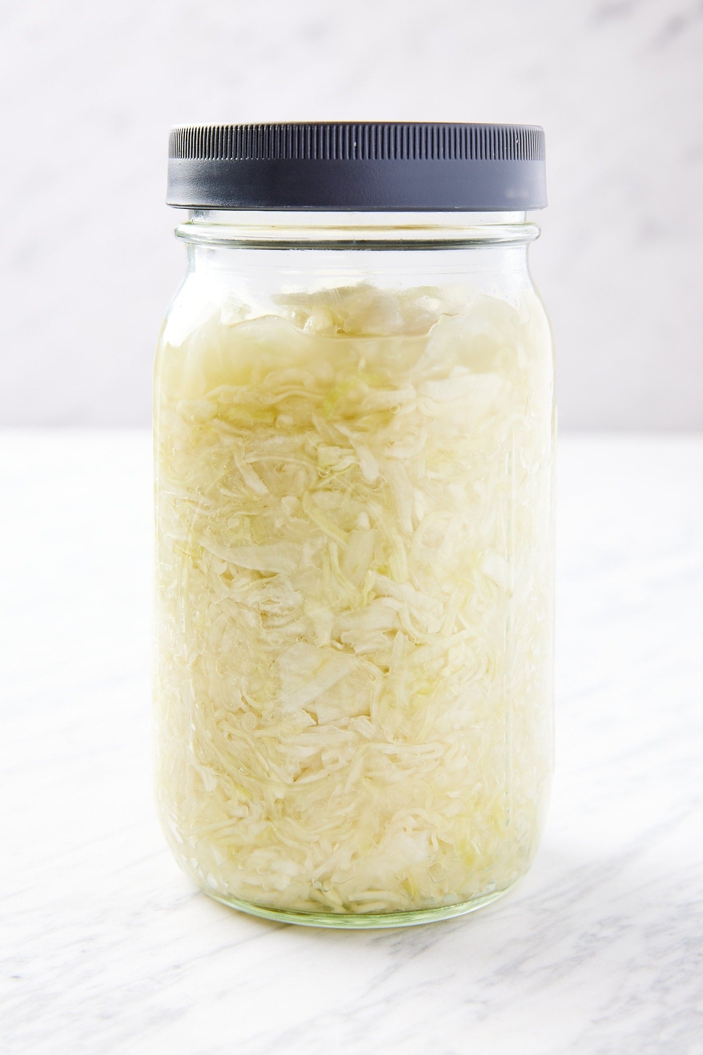 Quart jar with black screwtop lid filled with homemade sauerkraut sitting on white marble surface.