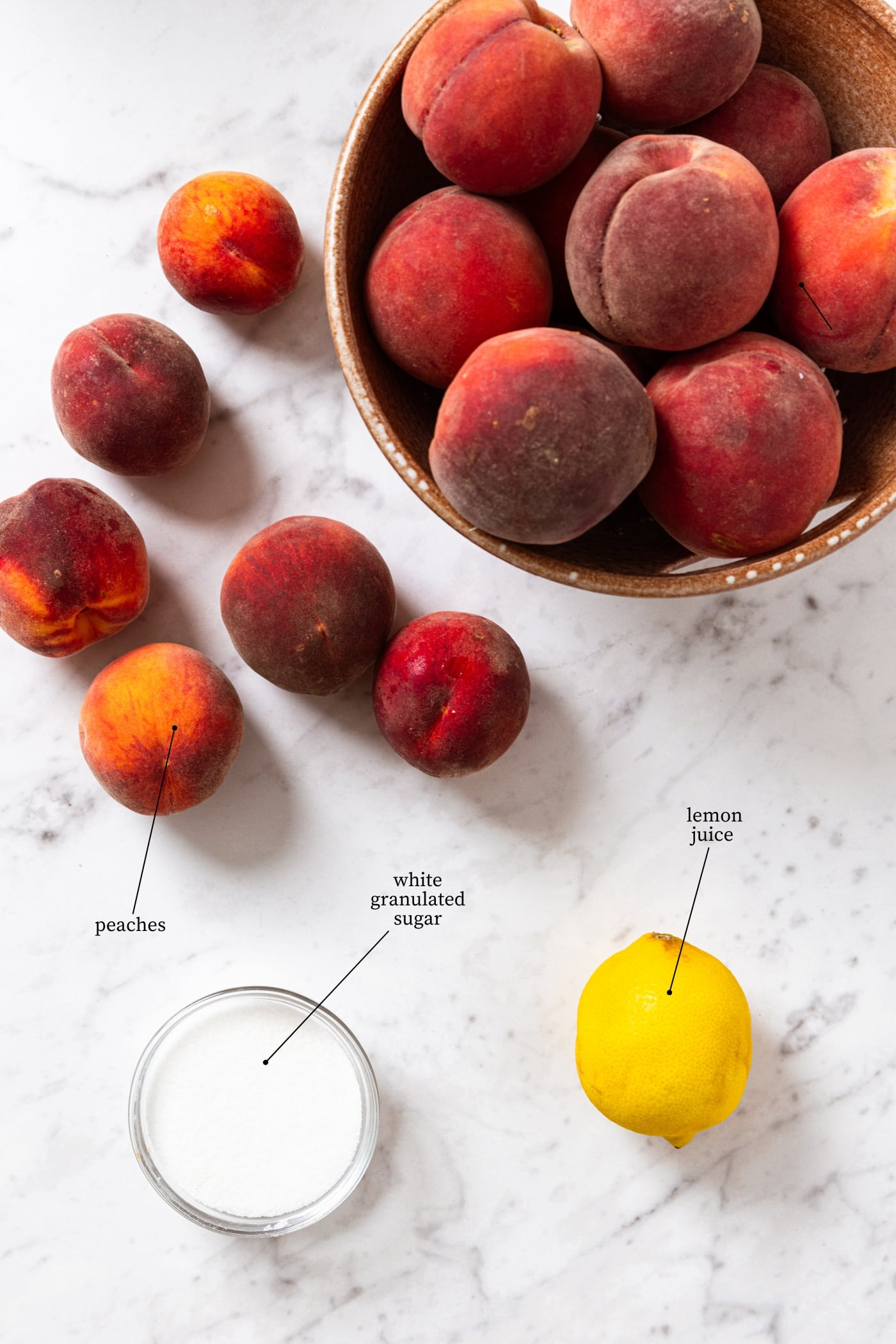 White marble surface filled with ingredients needed to make peach butter including peaches, lemon, and white sugar.