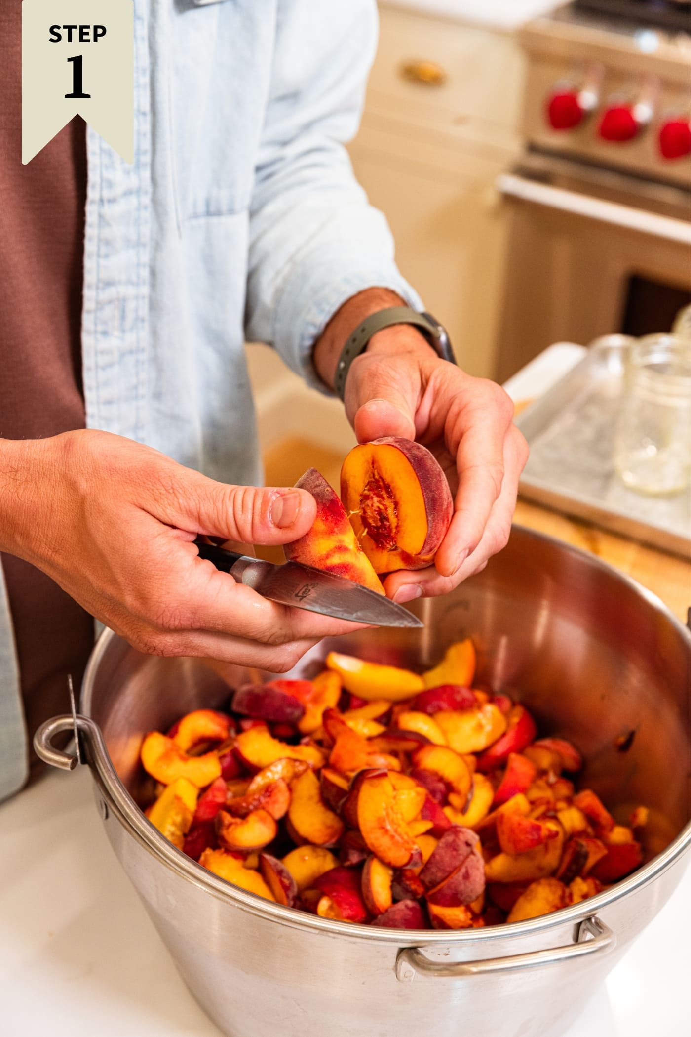 Two hands slicing apart a peach and taking out the inner pit with an entire pan of already sliced peaches sitting on white surface.