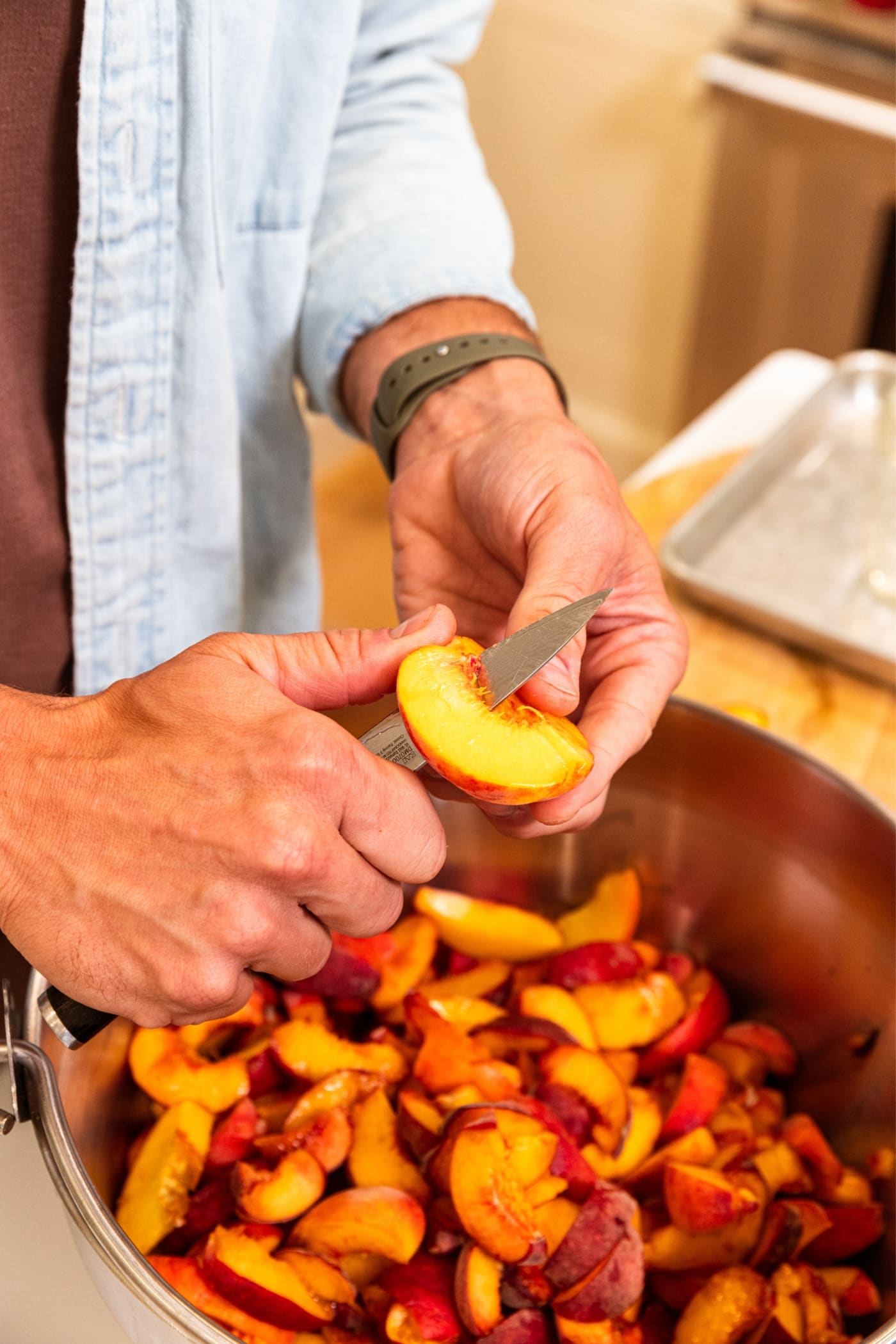 Hand using knife to slice peach into smaller slices above a pot already filled with lots of peaches.