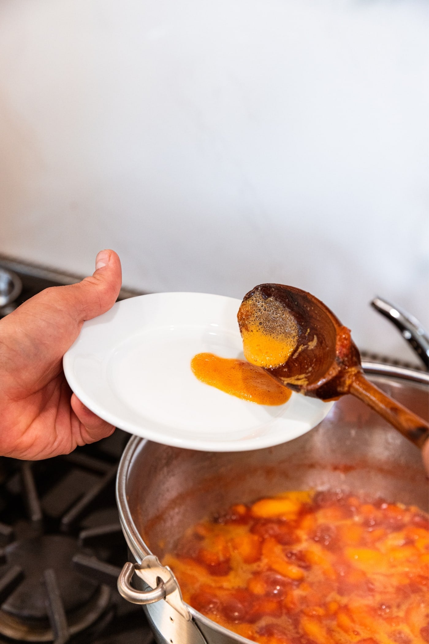 Hand holding small white plate scooping some of the foam from the top of cooking peach slices mixture in large pan on stovetop.