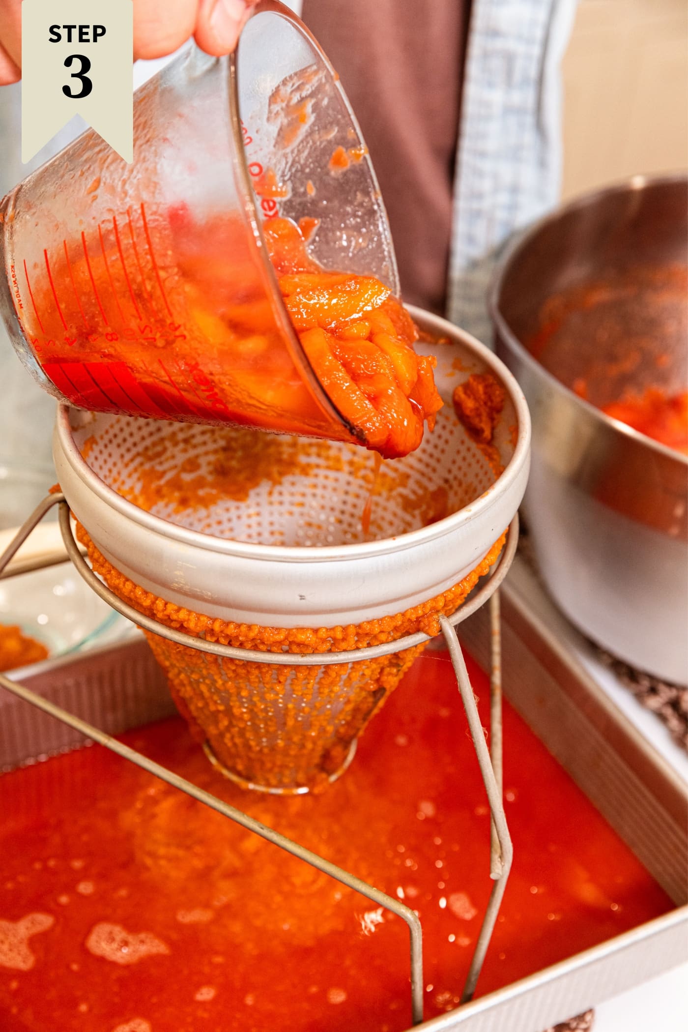 Hand holding glass measuring cup pouring peaches into a conical strainer set over a metal pan with peach juice leaking out.