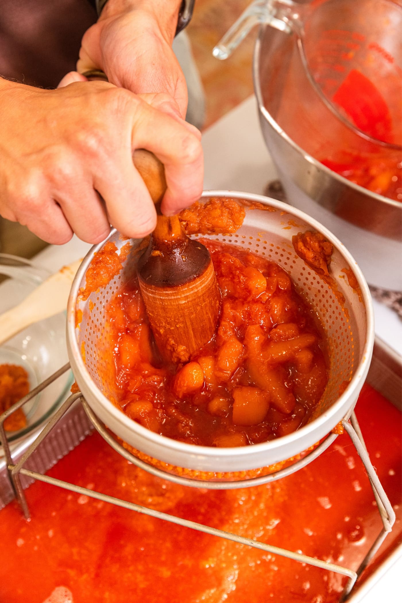 Hand using wooden roller to push bits of peach through the sides of a conical strainer into awaiting metal pan holding the peach juice.