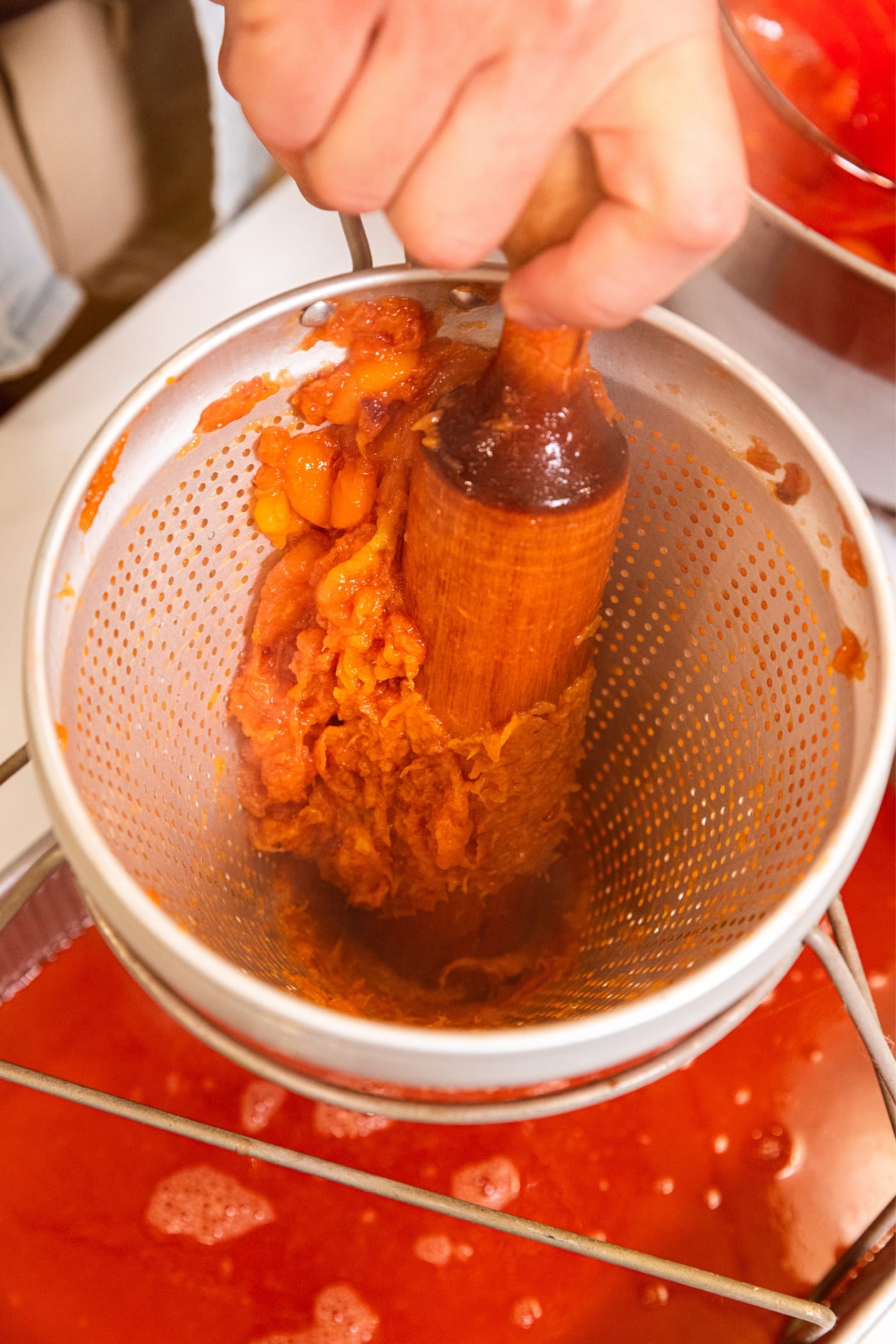 Hand holding wooden roller pressing bits of cooked peach through the sides of the conical strainer.