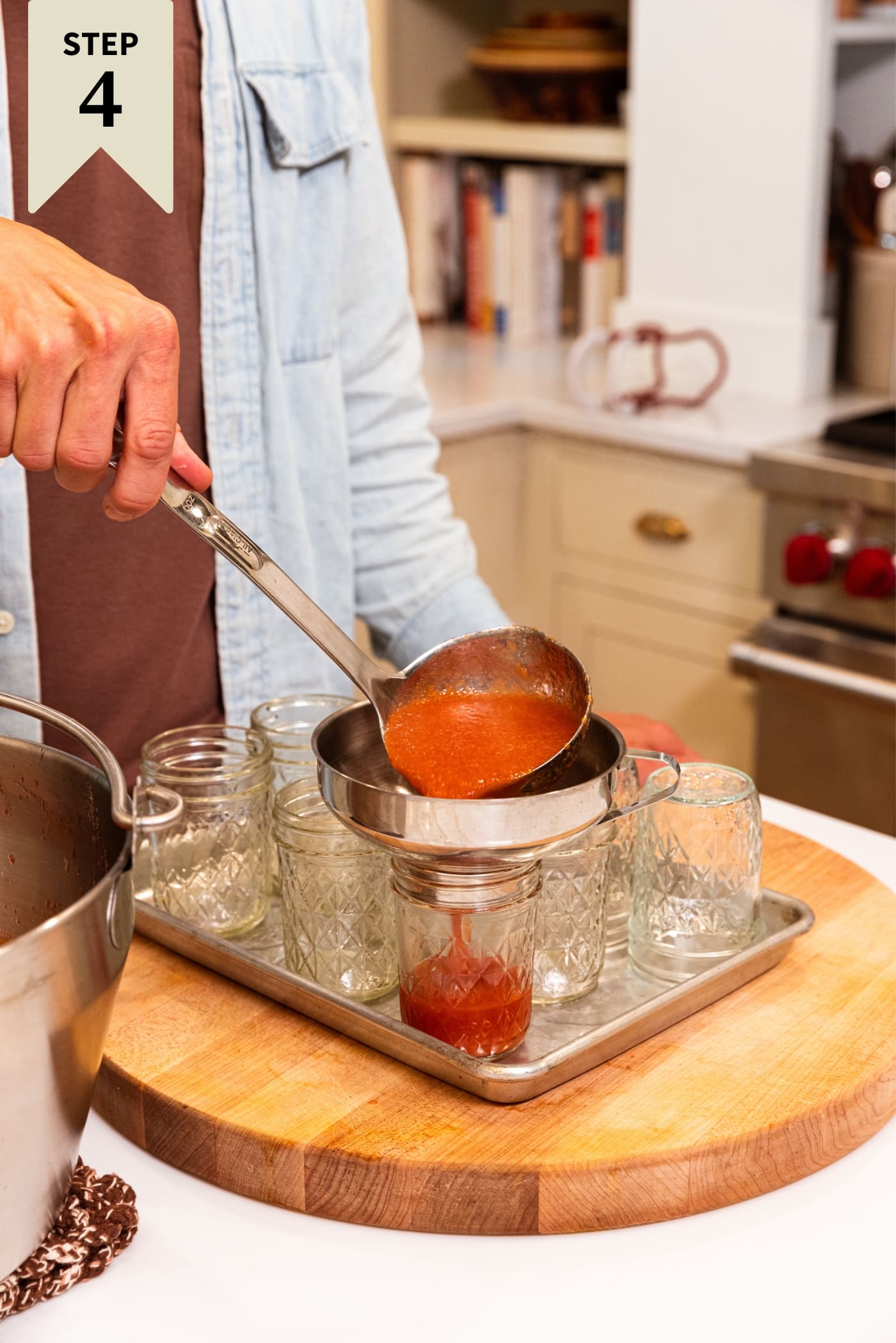 Hand holding ladle pouring peach butter into glass jam jars sitting on a metal baking pan.