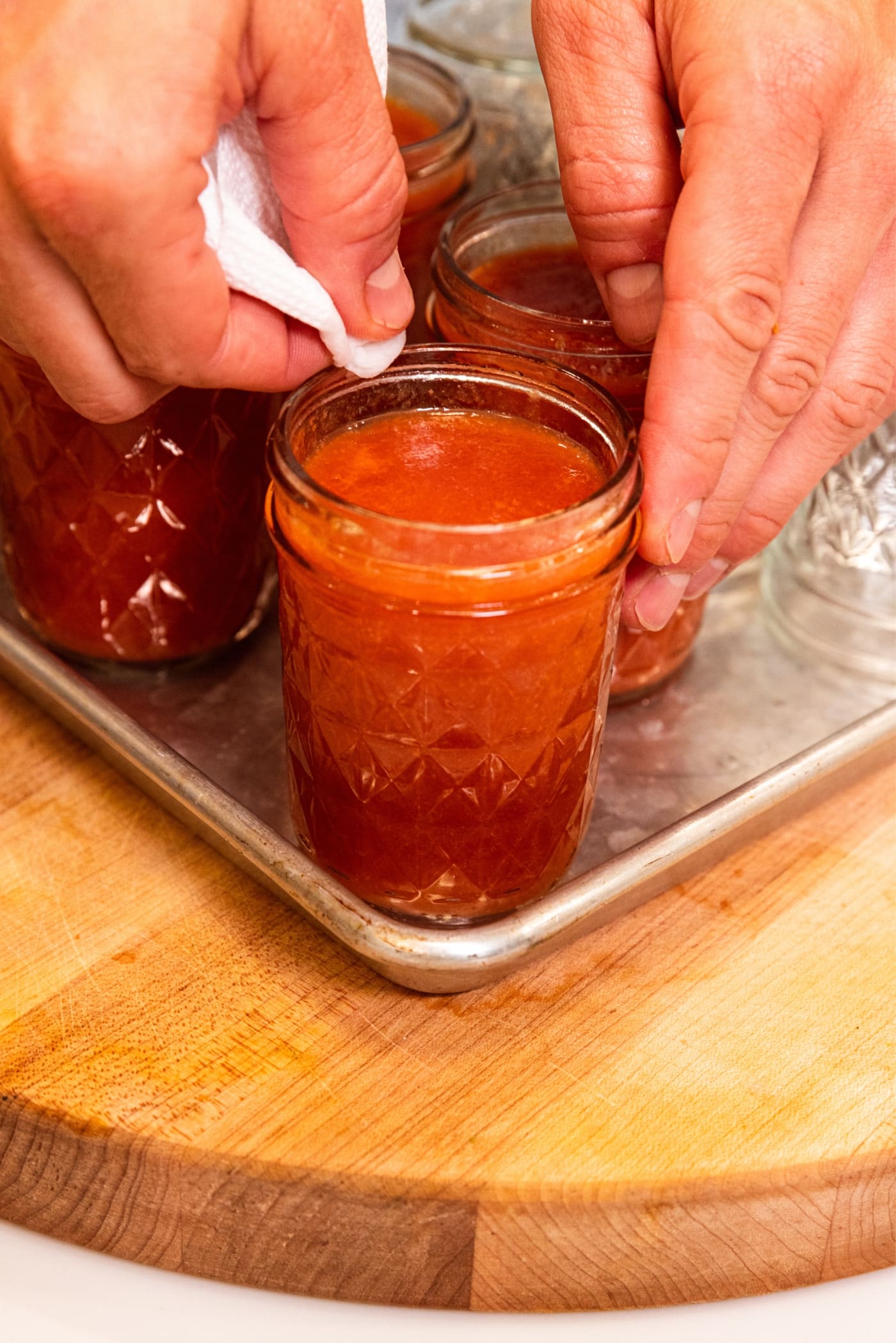 Hand wiping off the rim of a glass jam jar filled with peach butter sitting on metal baking sheet.