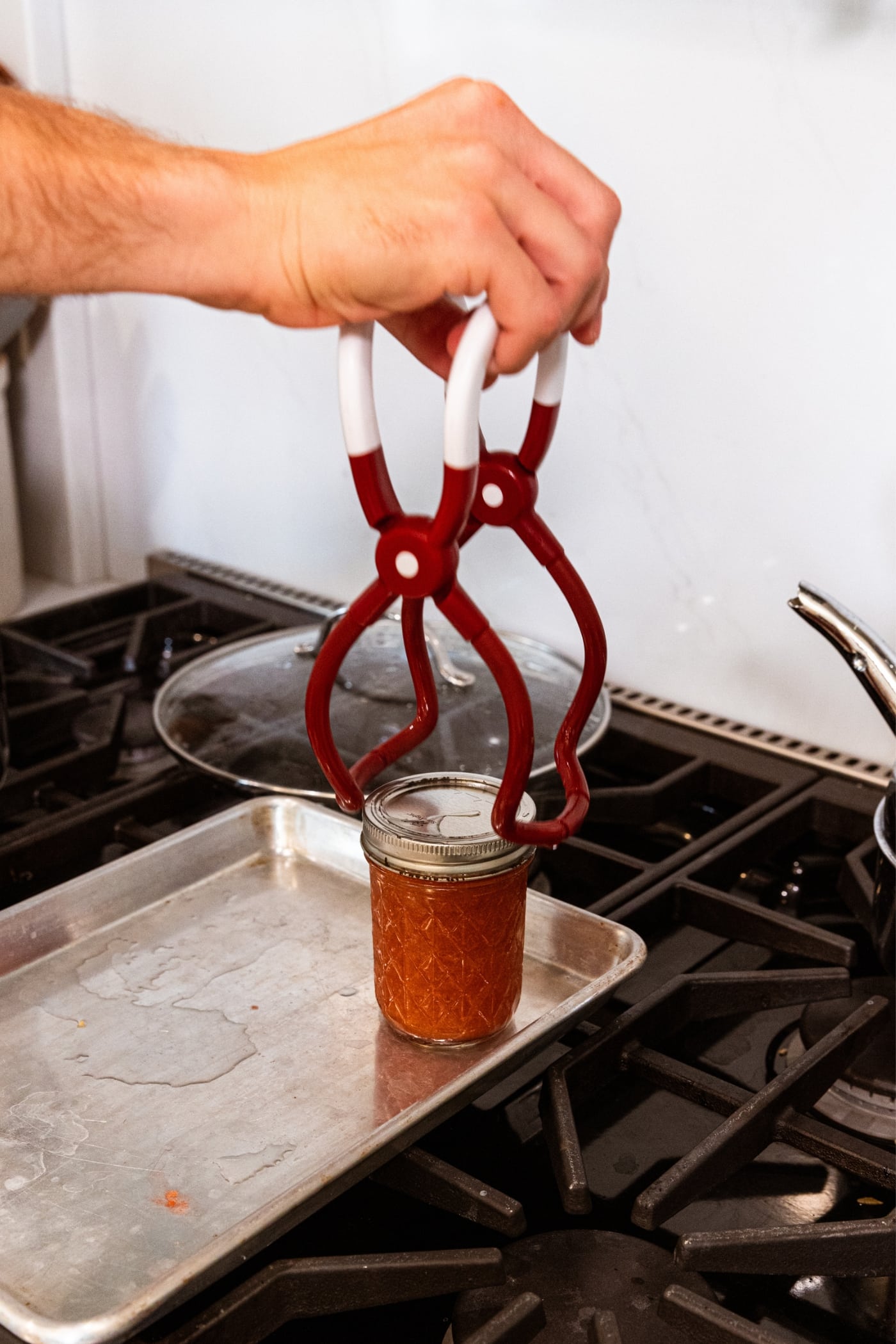 Hand holding jar lifter putting processed jam jar filled with peach butter onto a metal baking sheet.