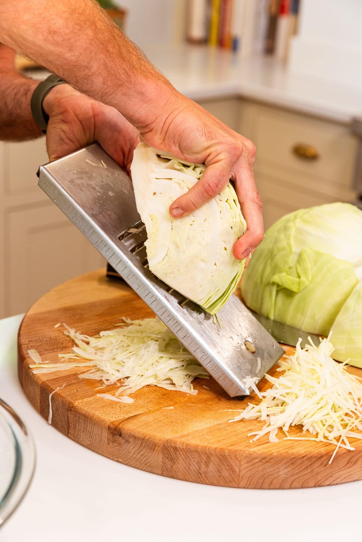 Hand holding mandolin slicing quarter of a head of cabbage with shreds of cabbage underneath all on wooden cutting board.