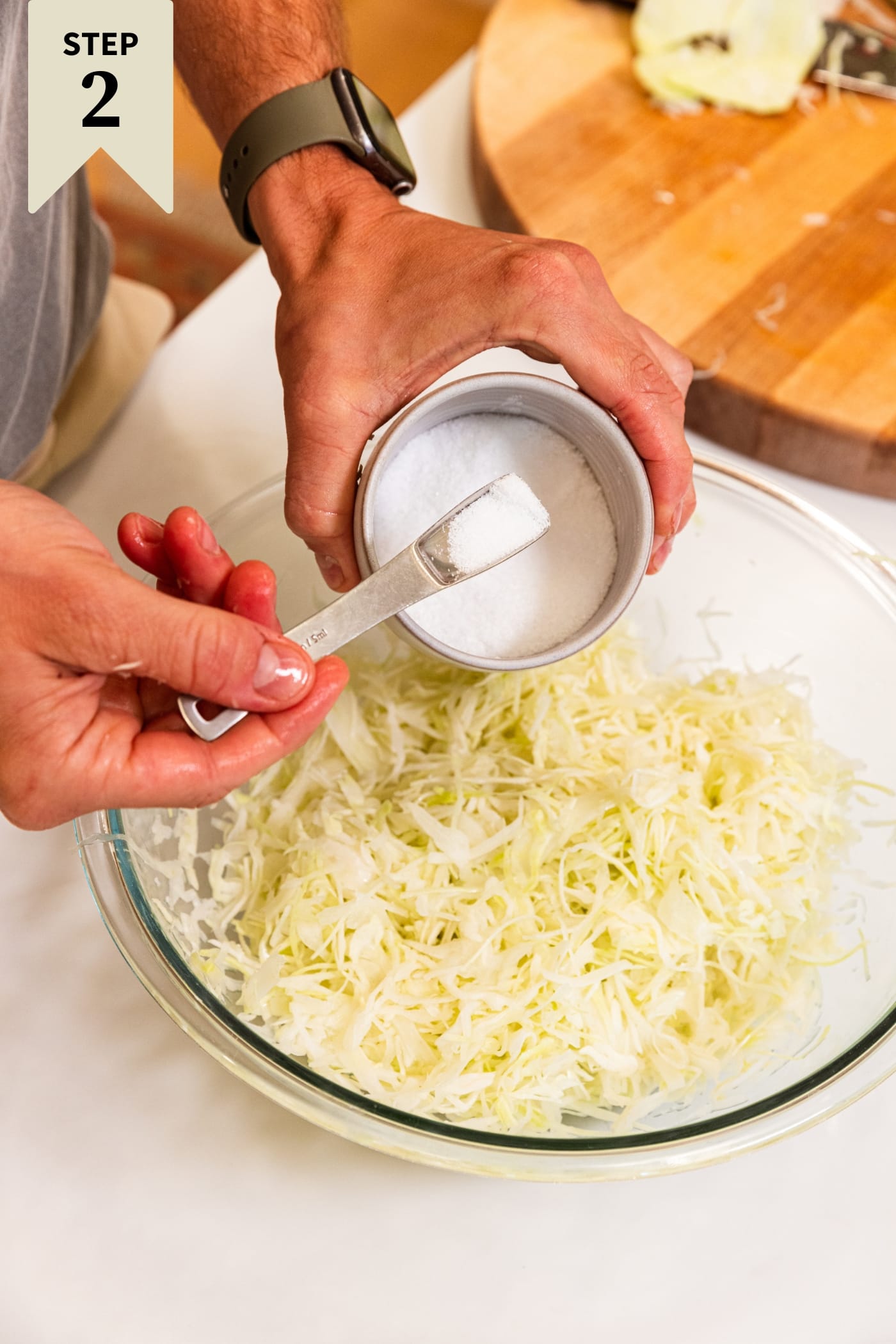 Hand holding teaspoon filled with kosher salt from salt cellar about to sprinkle it on top of shredded cabbage in a large glass bowl.