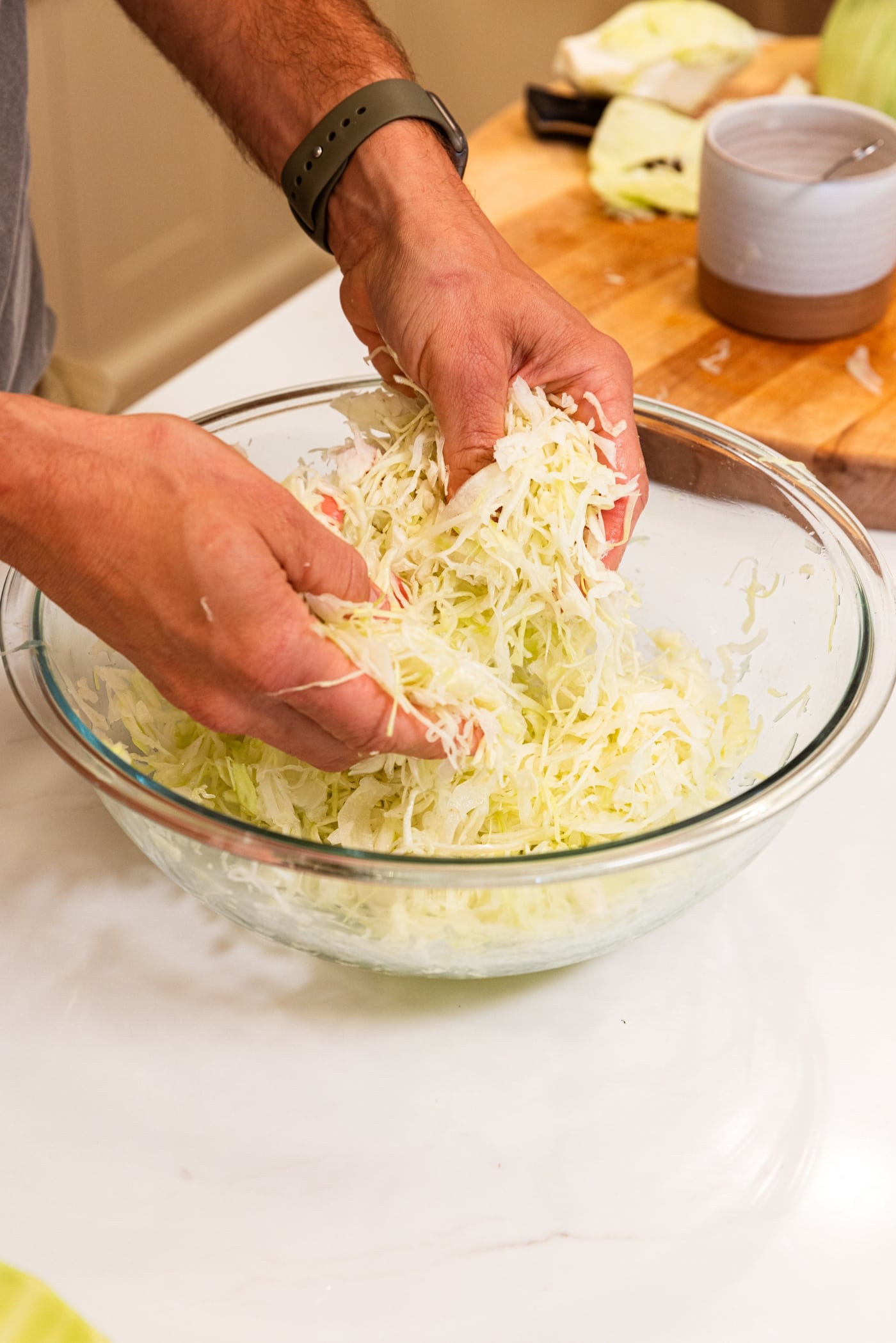 Two hands squishing shredded cabbage inside of glass bowl after spreading kosher salt all over the top.