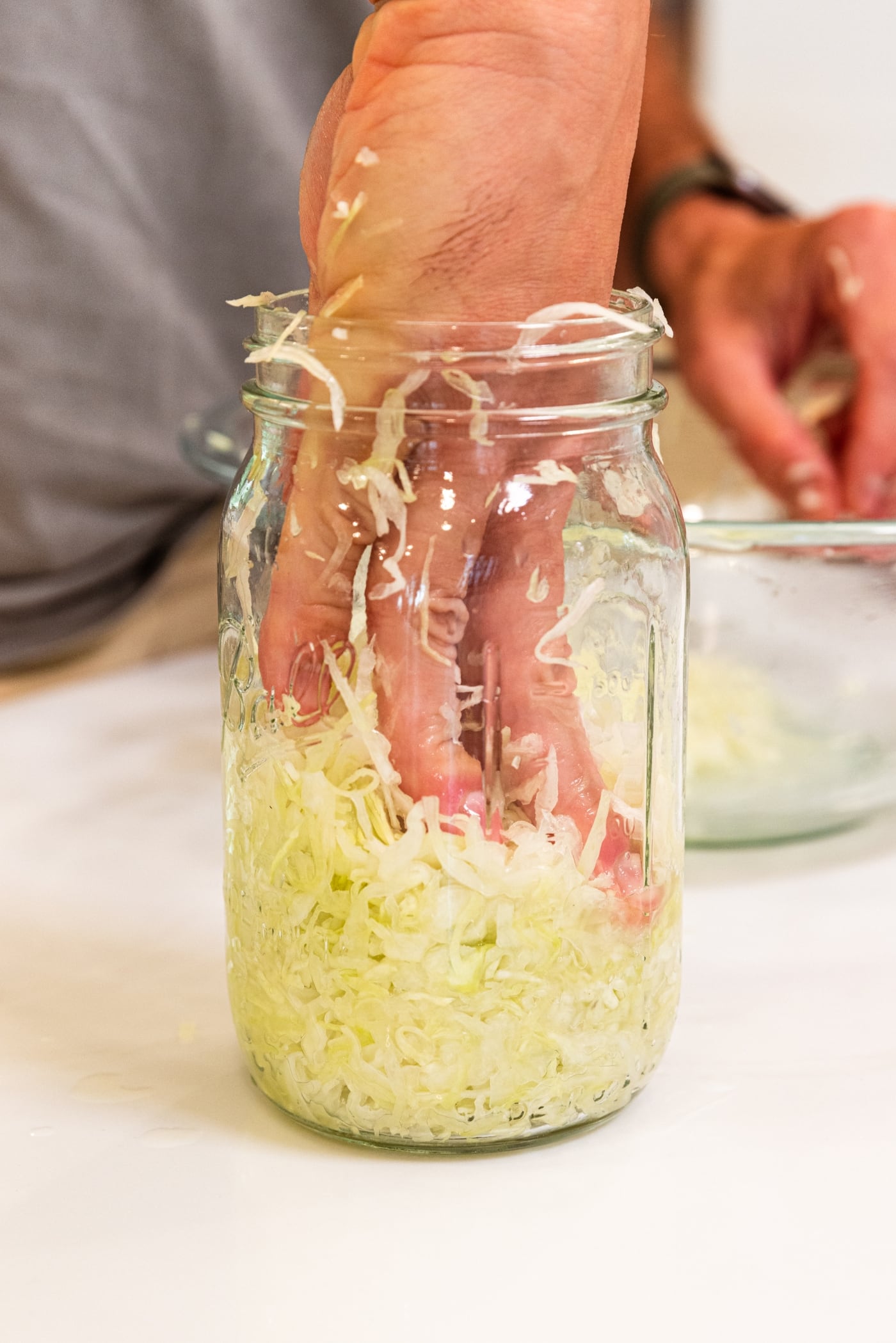 Hand using fingers to press shredded cabbage into a quart jar on white surface and get the juices to exude.