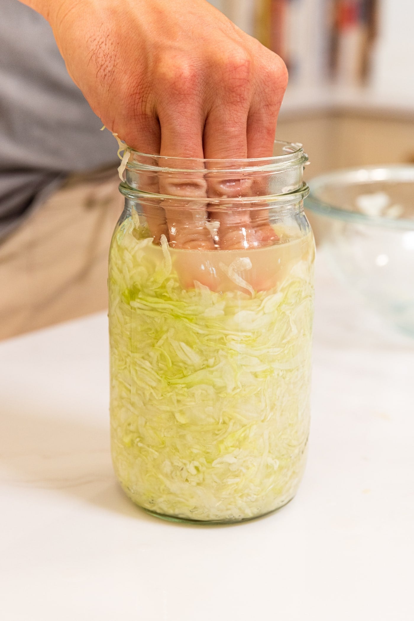 Hand pressing shredded cabbage into a quart jar to make homemade sauerkraut all on white surface in kitchen.