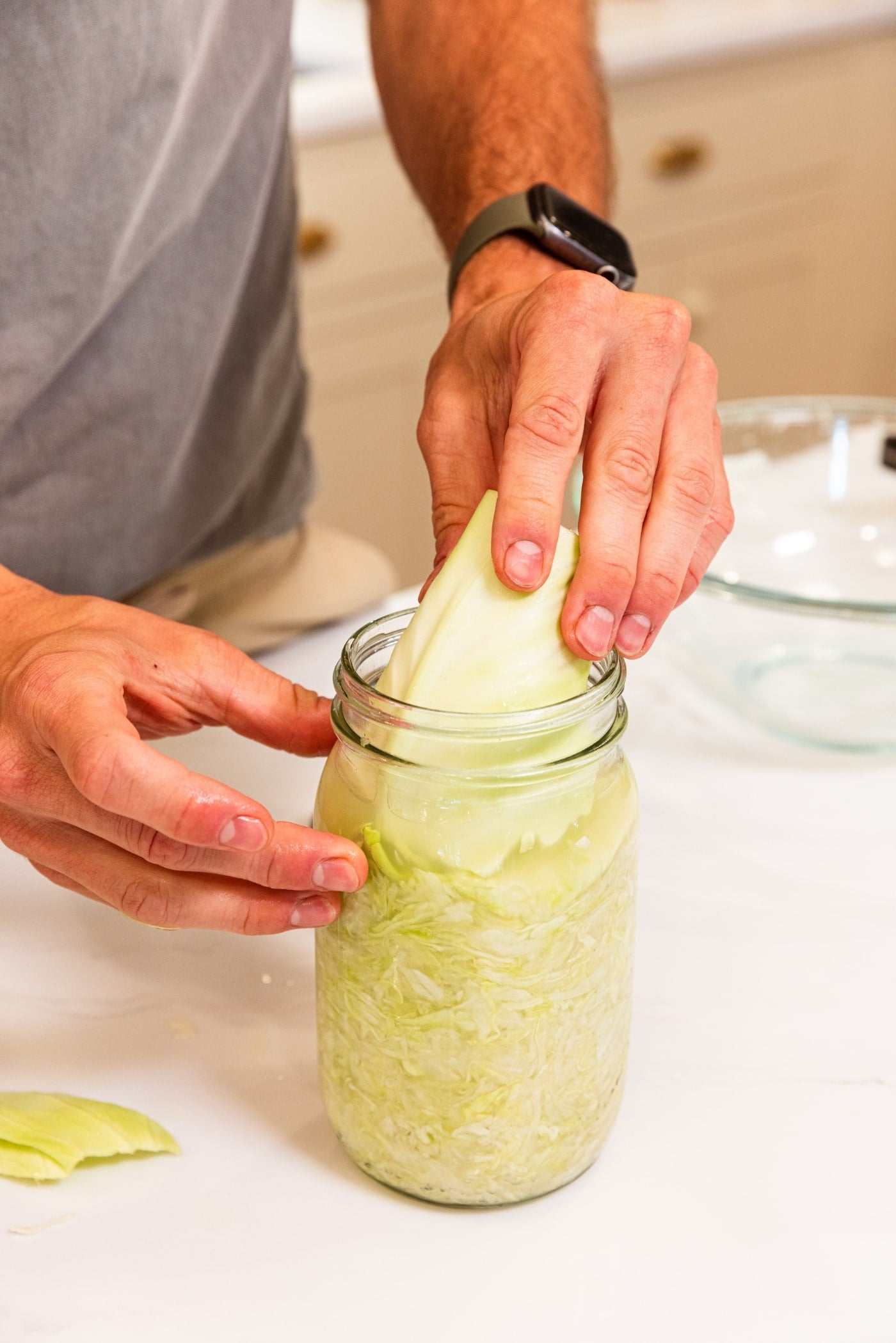 Two hands pressing hard piece of cabbage into a quart jar filled with fermenting sauerkraut on white surface.