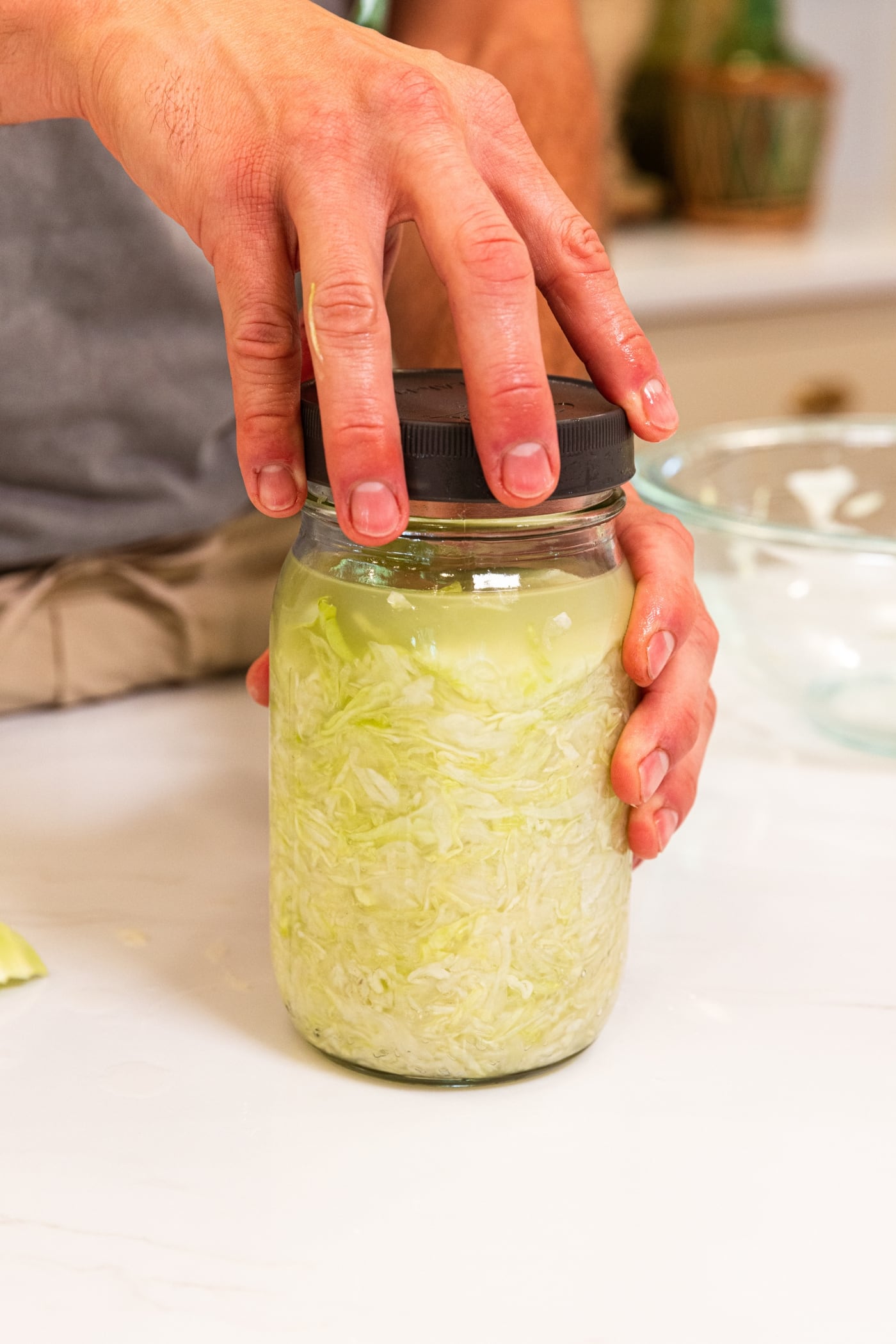 Hand holding lid screwing it onto the top of a quart jar filled with fermenting sauerkraut on a white surface.