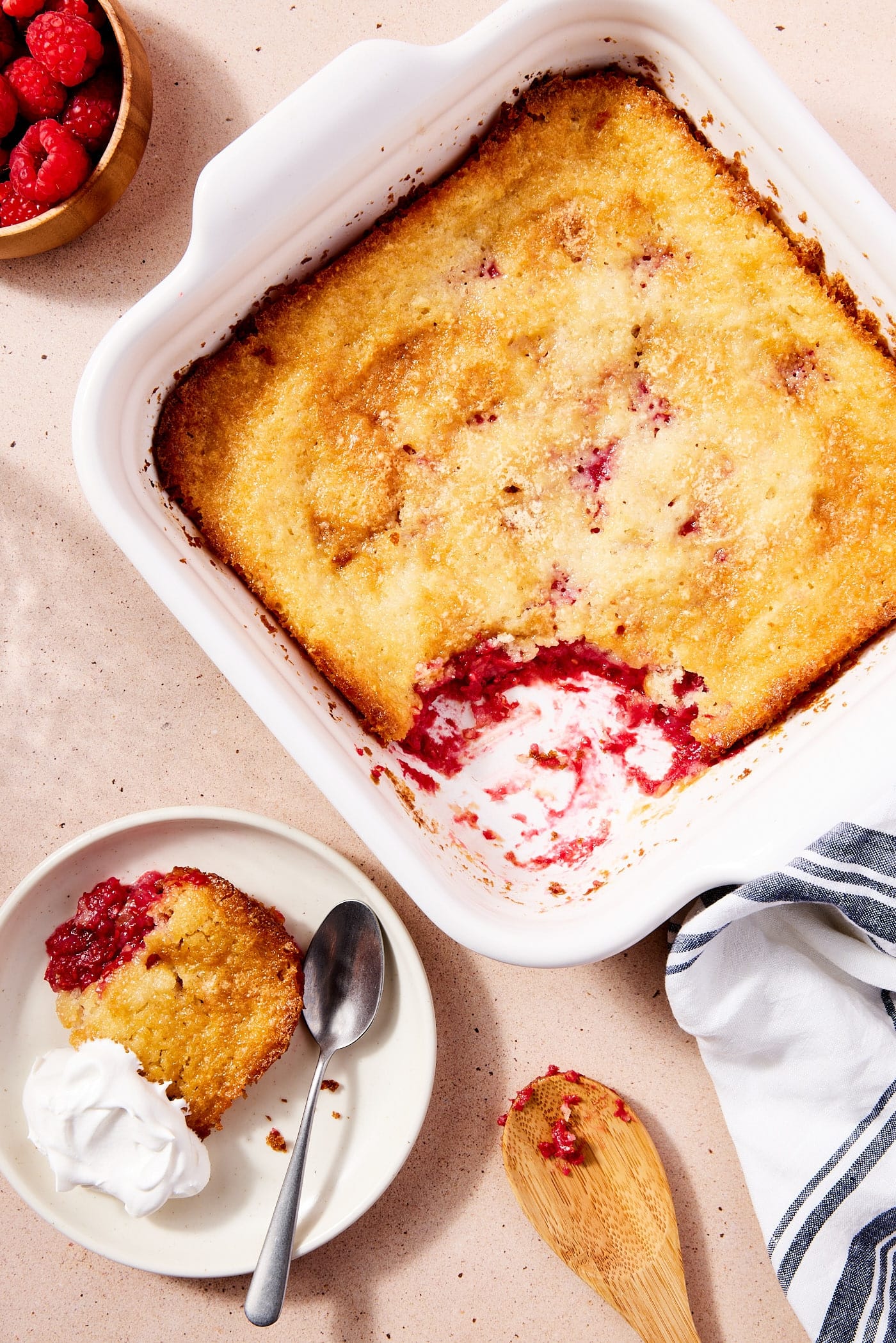 Baked lazy raspberry cobbler in white baking dish with scoop taken out and placed on white plate with a dollop of whipped cream.