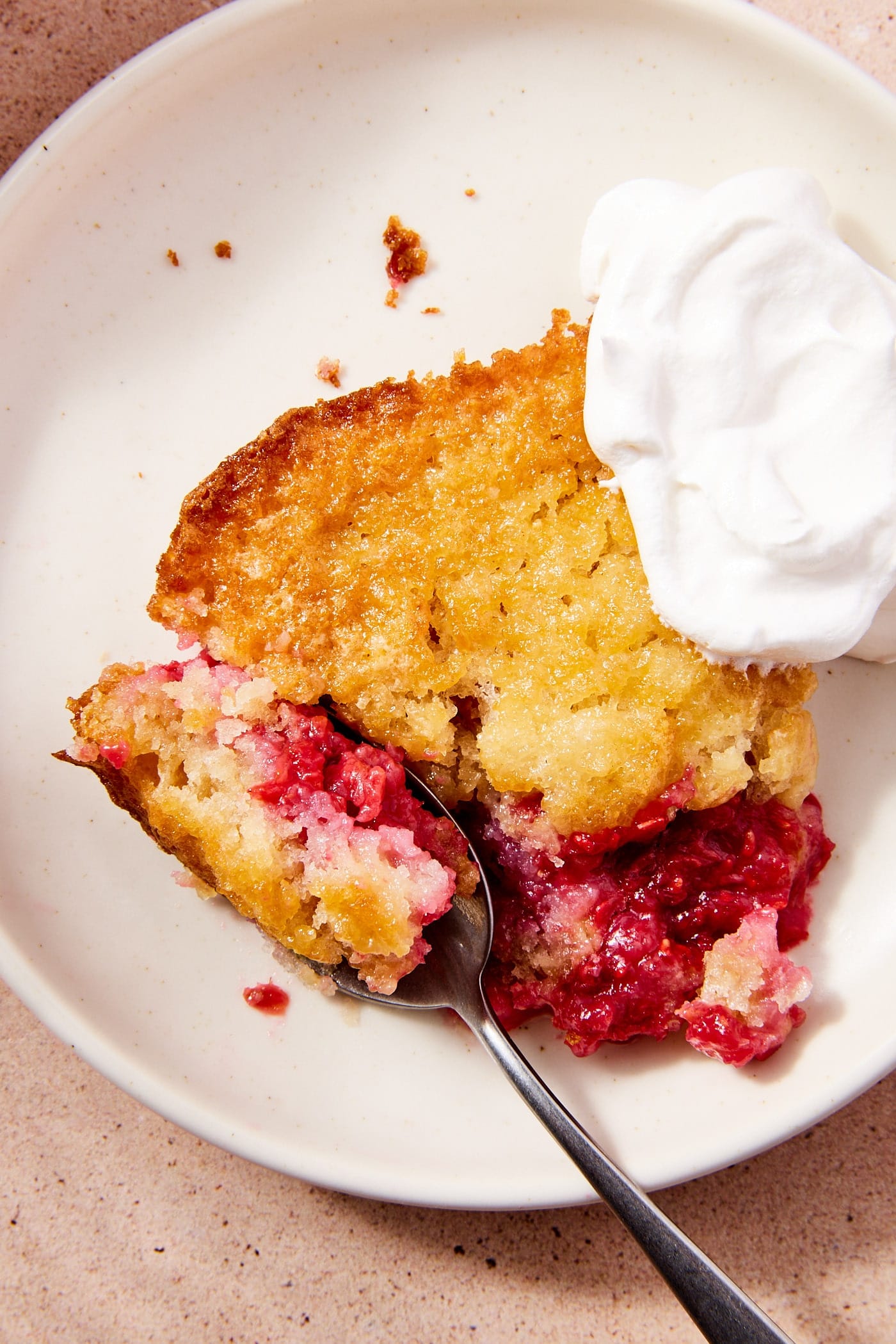 White plate with scoop of raspberry cobbler with spoon taking a piece from the slice along with dollop of whipped cream to the side.