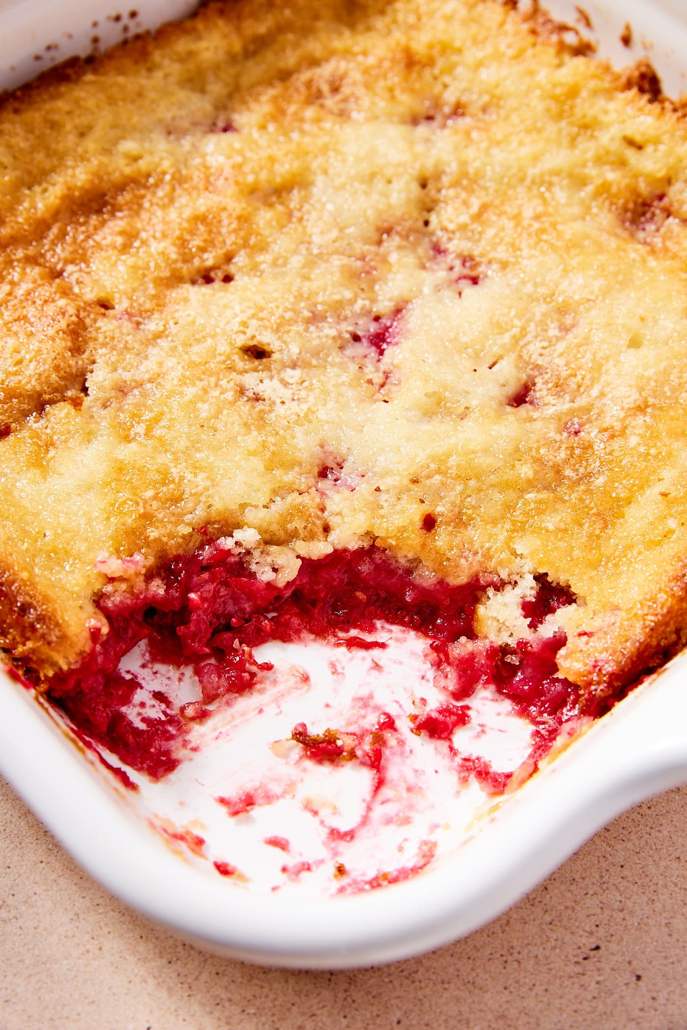 White baking dish filled with baked raspberry cobbler with scoop removed showing interior red filling with cobbler on the top.