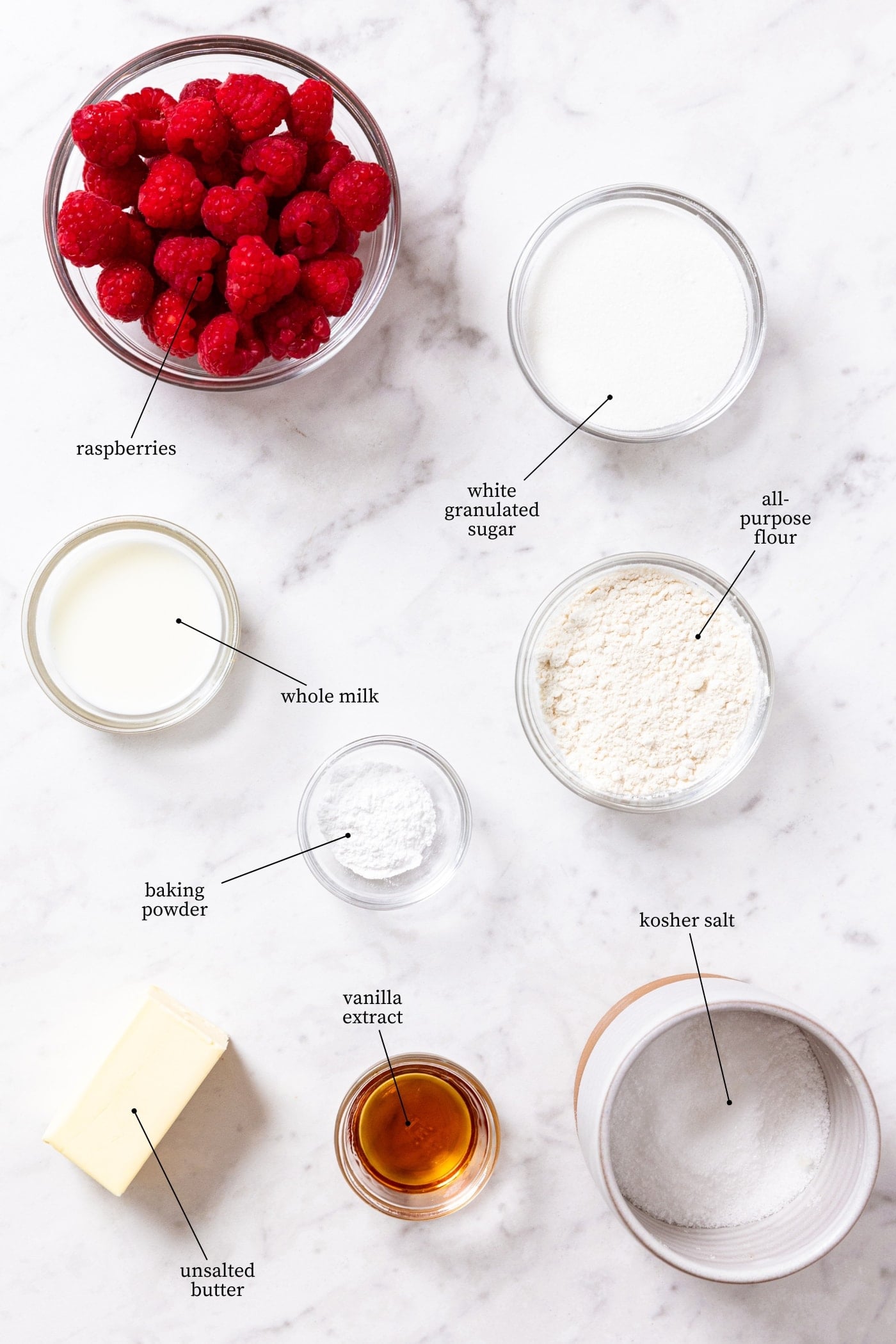 White marble surface filled with all ingredients needed to make a lazy raspberry cobbler including flour, sugar, butter, vanilla, raspberries, and more.