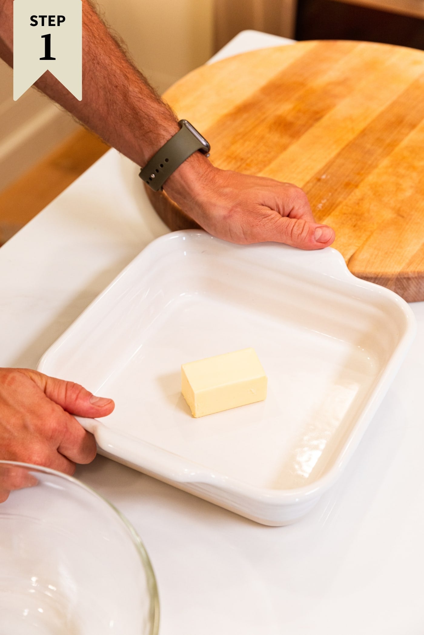 Hand holding onto the sides of a white baking dish with half a stick of butter resting in the bottom of the pan before being melted in the oven.