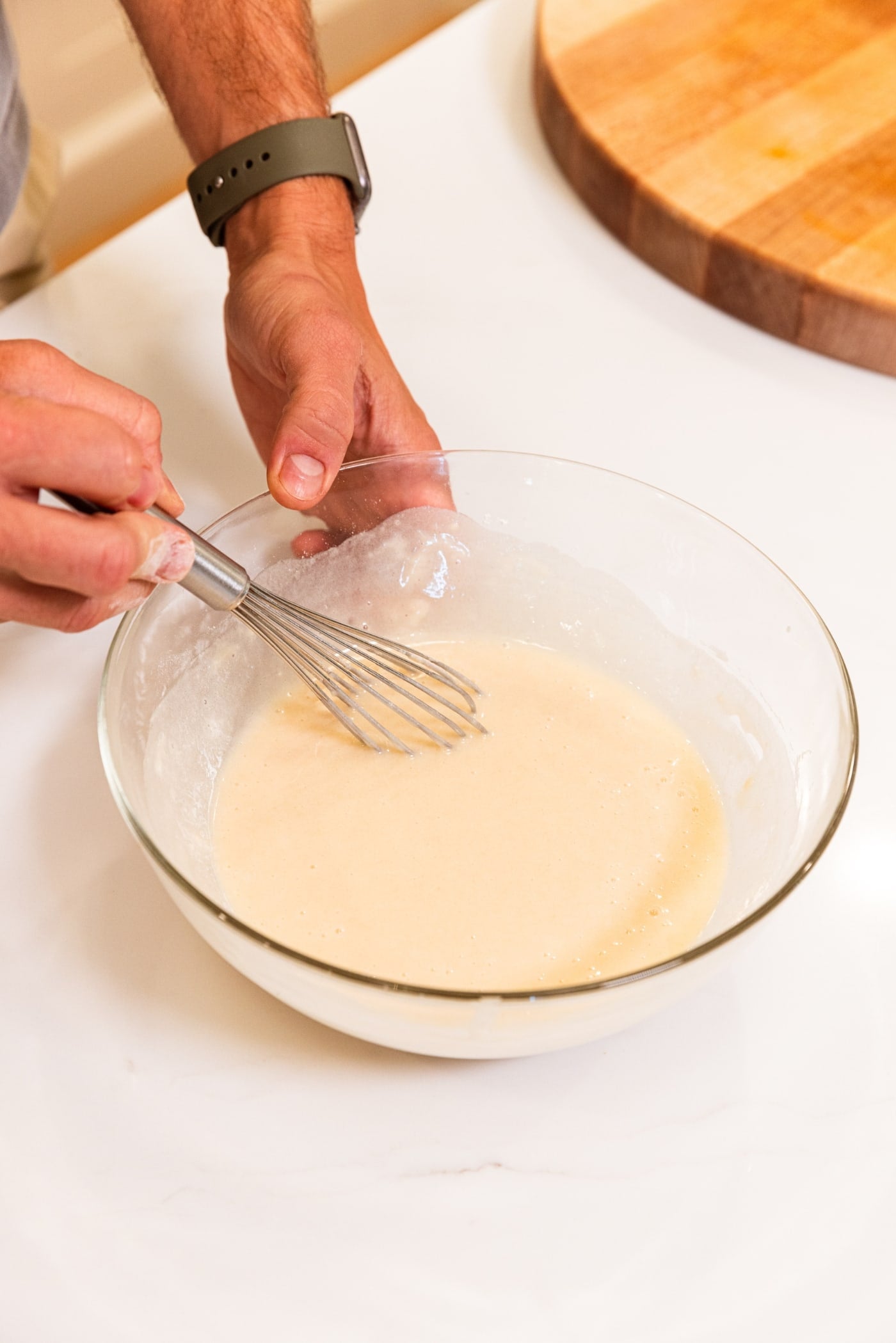 Hand holding glass mixing bowl whisking together ingredients for a cobbler batter all on white surface.