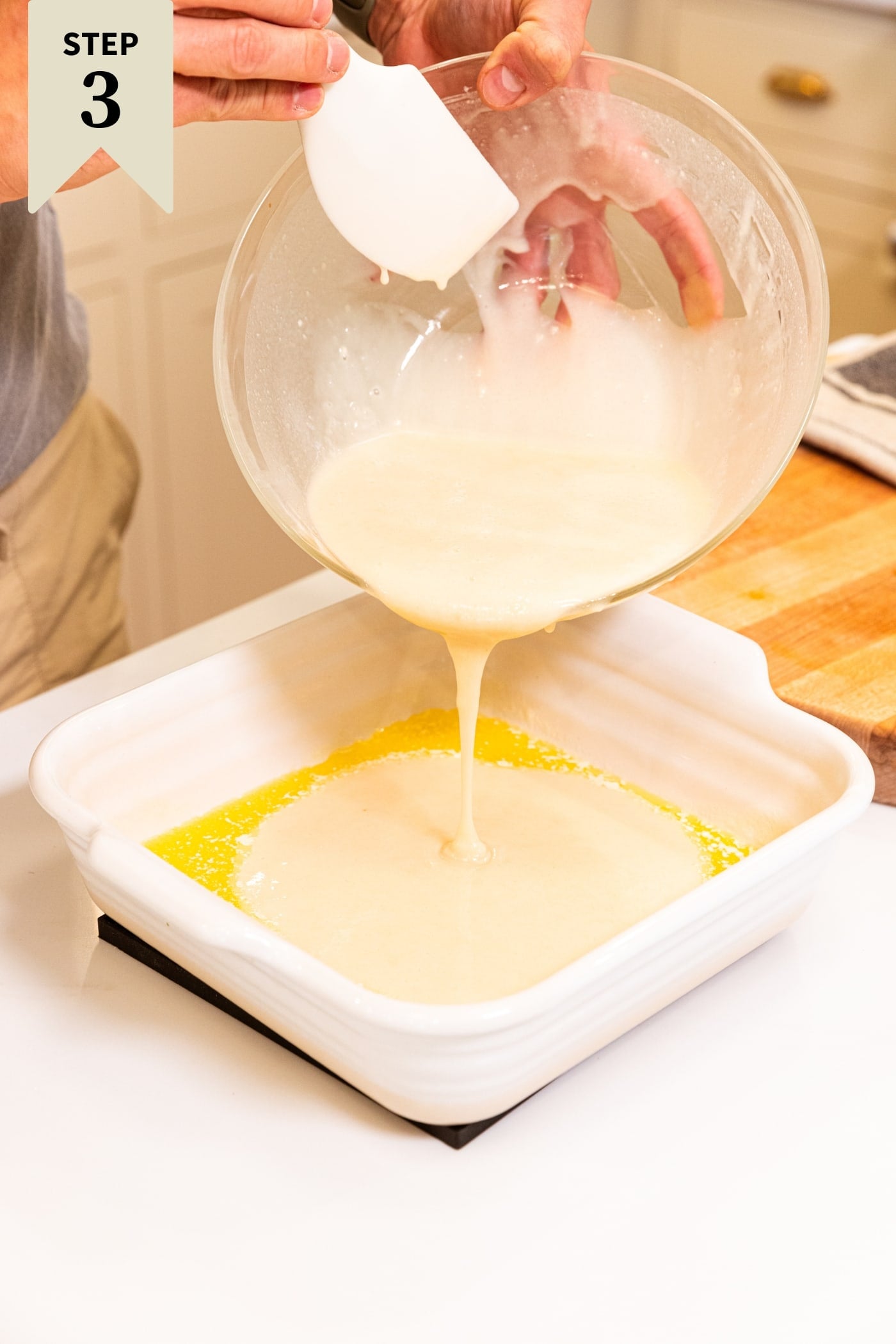 Hand holding glass bowl pouring batter on top of melted butter in white baking dish.
