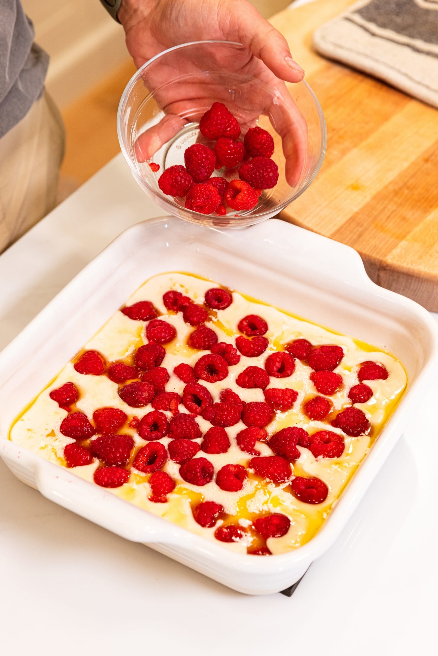 Hand holding small glass bowl pouring raspberries on top of batter in white baking dish.