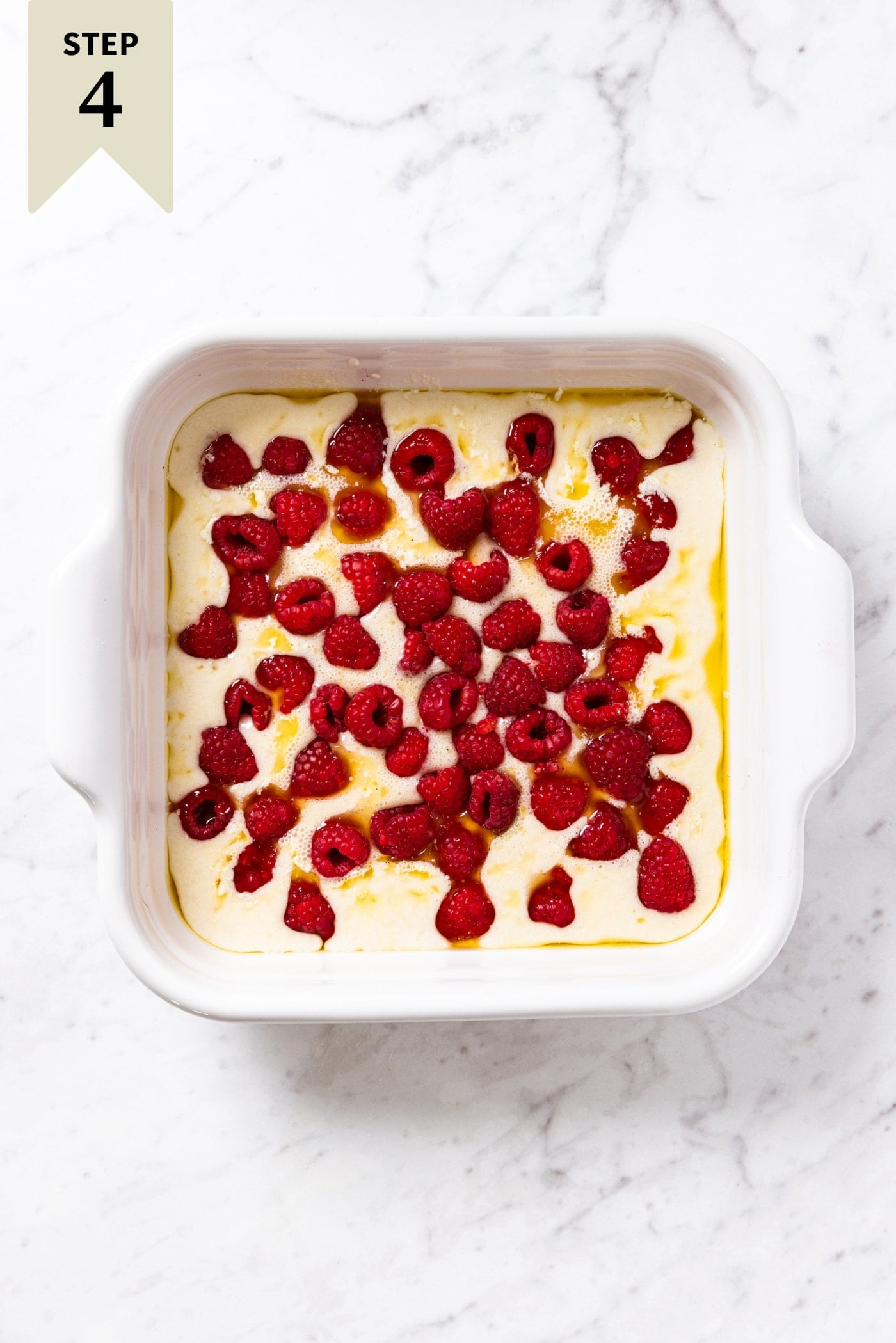 Unbaked lazy raspberry cobbler sitting in white baking dish on white marble surface with raspberries all over the top.
