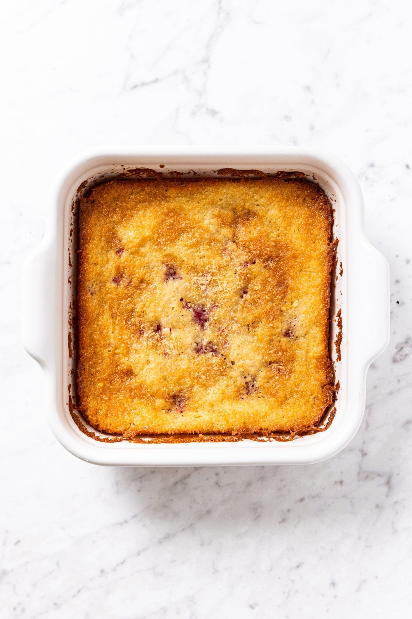 White marble surface with white baking dish filled with baked raspberry cobbler with small parts of raspberries peeking through.