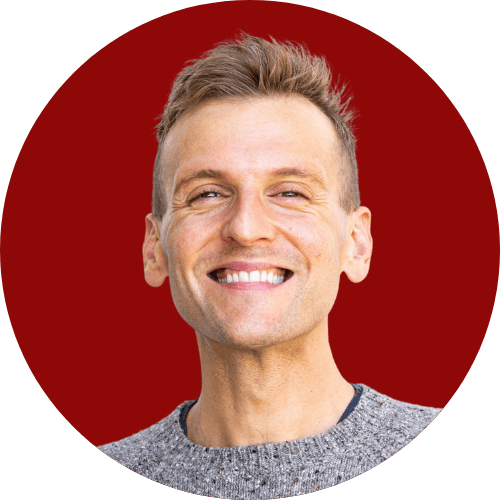 Kaleb Wyse wearing gray sweater standing in front of red background smiling.