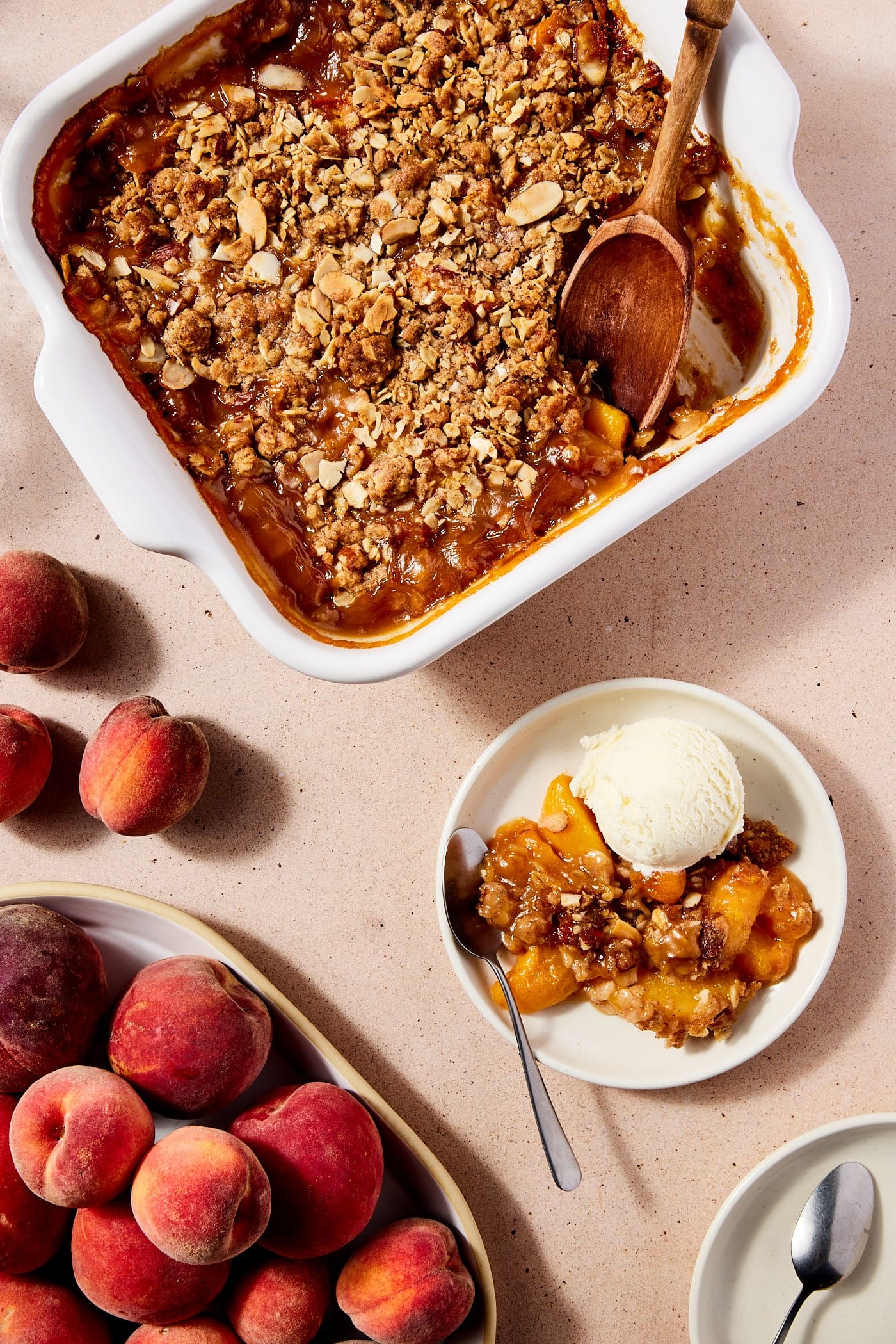 Light pink surface filled with baked peach crisp with a scoop sitting on a plate with a scoop of ice cream as well and peaches.