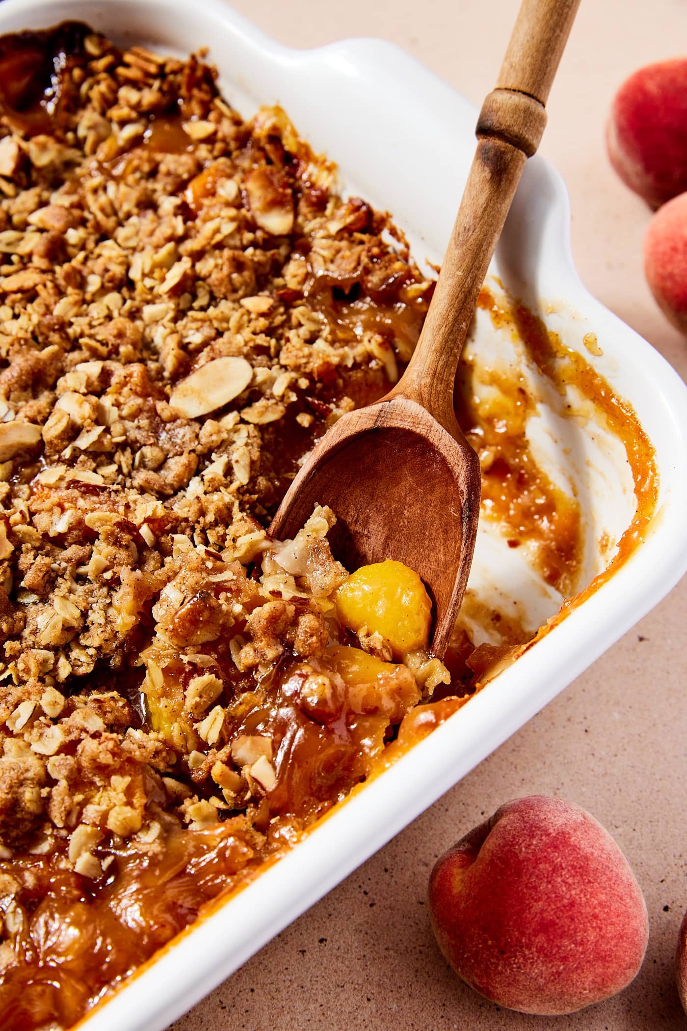 White baking dish filled with baked peach crisp with streusel topping with one piece of peach sticking out on wooden spoon.