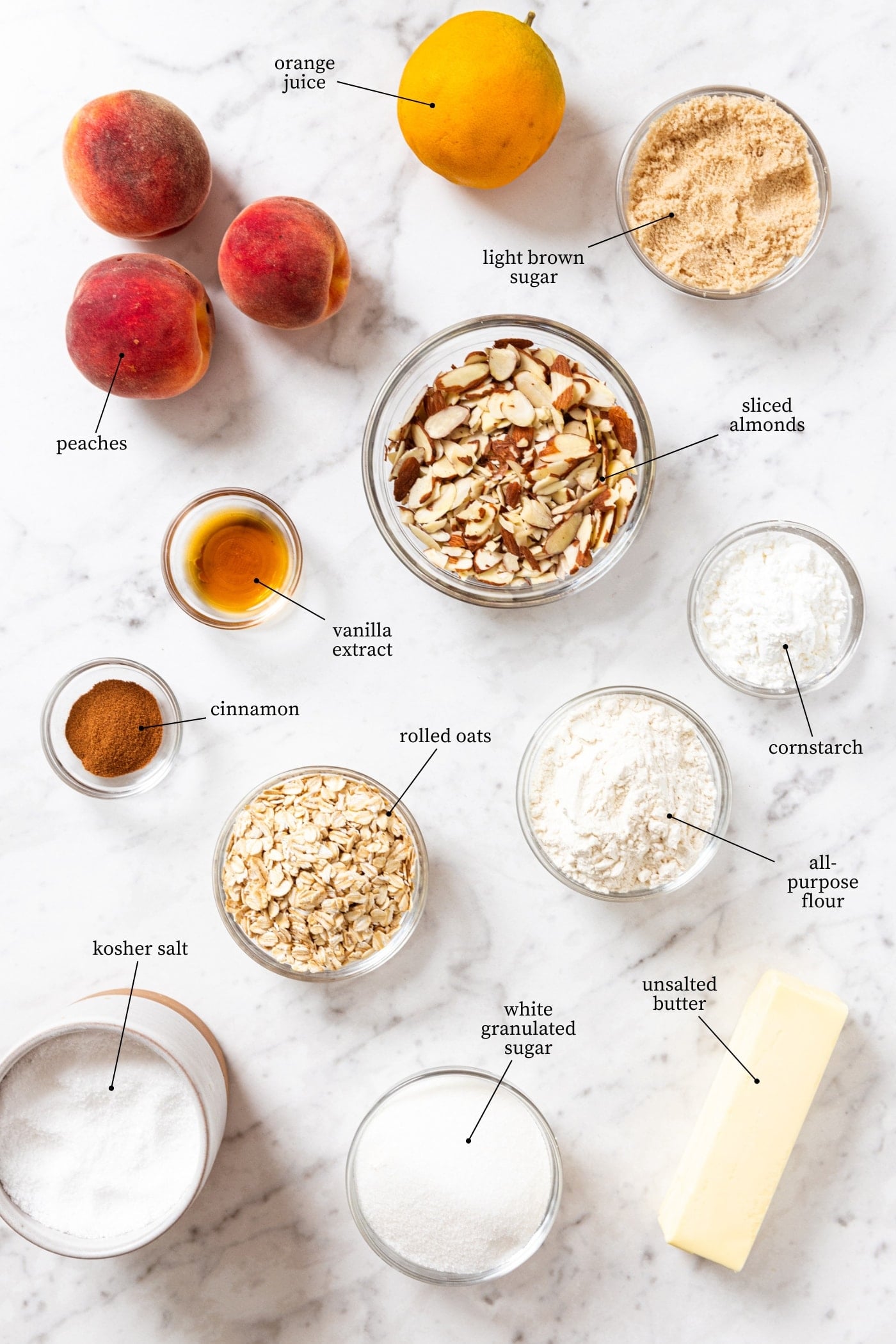 White marble surface filled with all ingredients needed to make peach crisp including peaches, sugar, flour, almonds, oats, butter, and more.