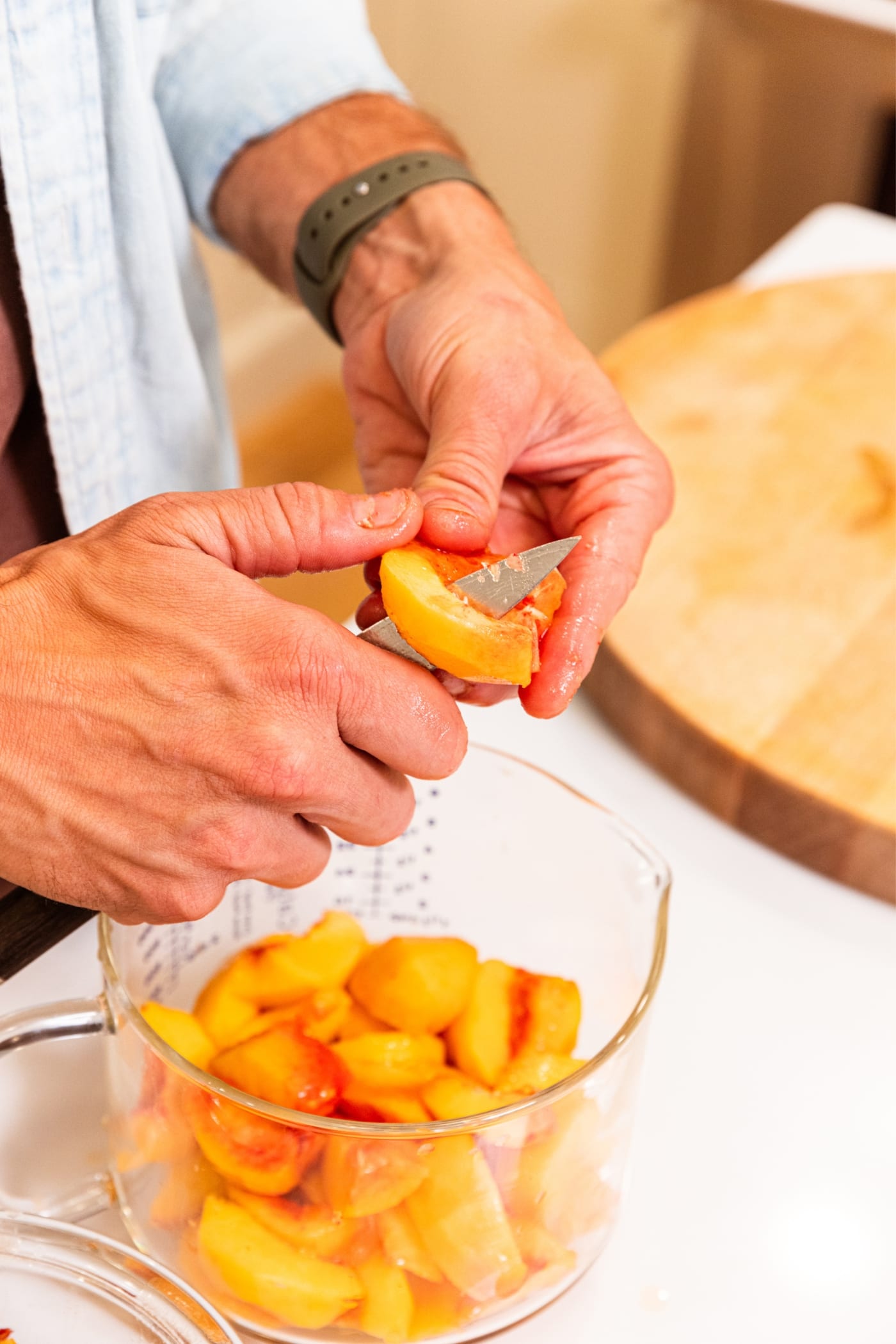 Hand using knife to slice a peach into smaller slices with measuring cup below containing the peach slices.