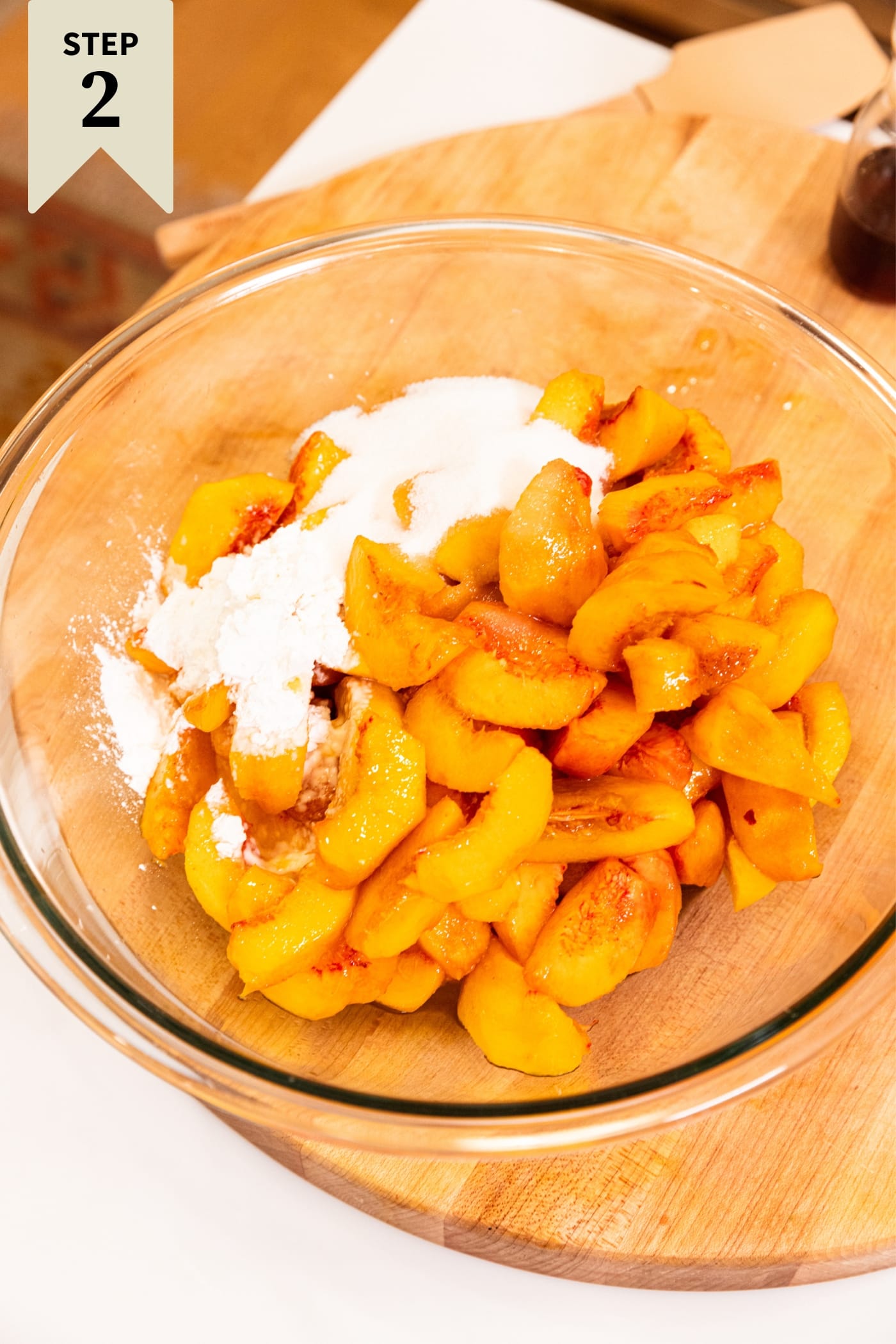 Large glass bowl filled with slices of peach slices with sugar, flour, and spices in the bowl sitting on wood cutting board.