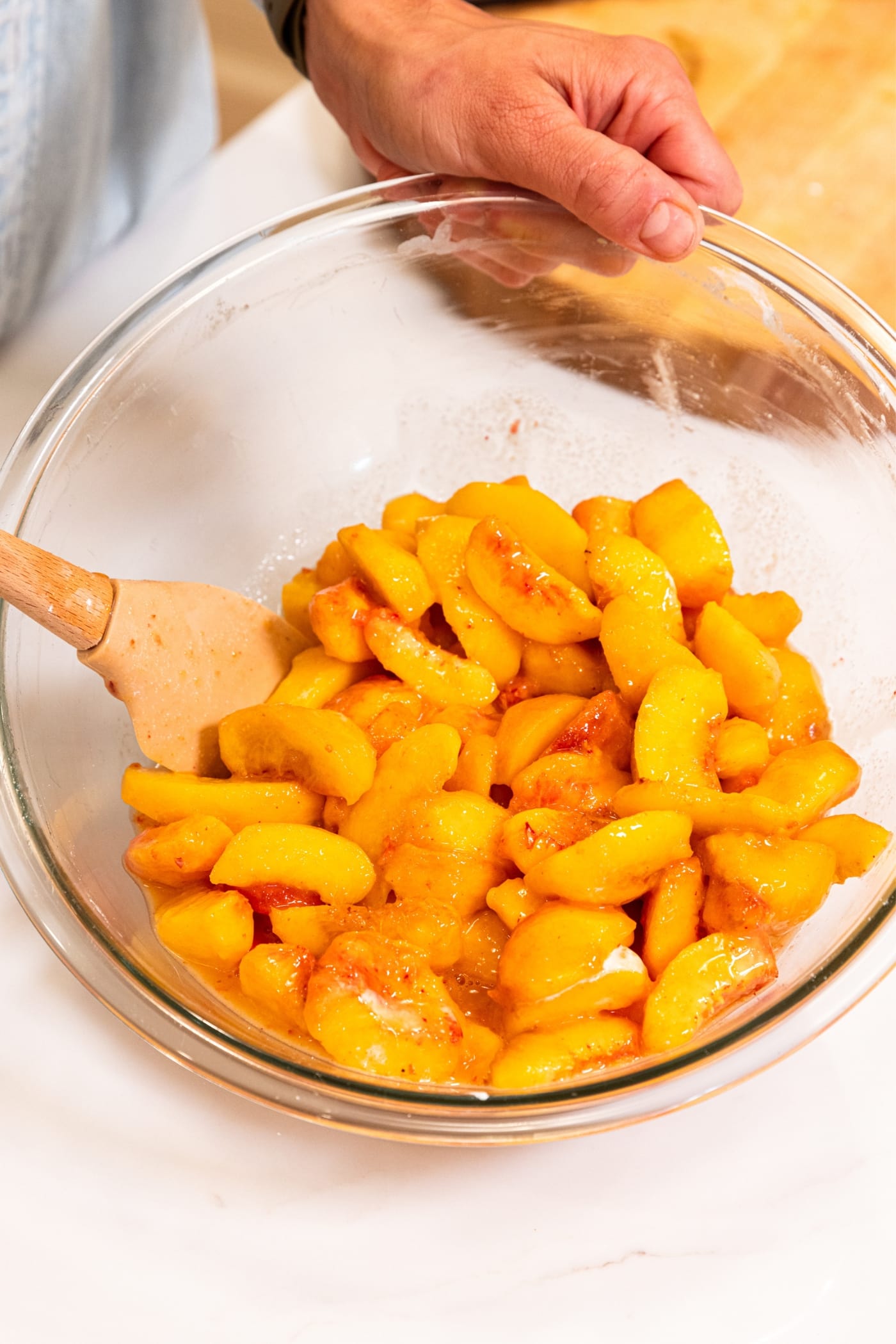 Hand stirring together peach mixture with plastic spatula in glass mixing bowl on white surface.