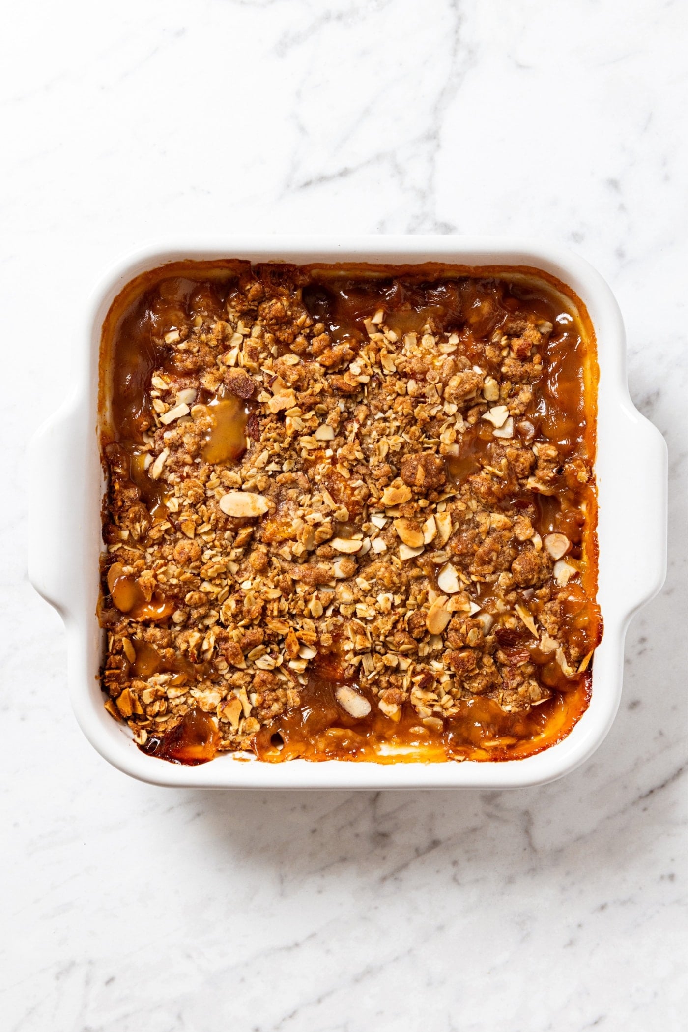 Baked peach crisp sitting on white marble surface after coming out of the oven with bumbling around the edges.