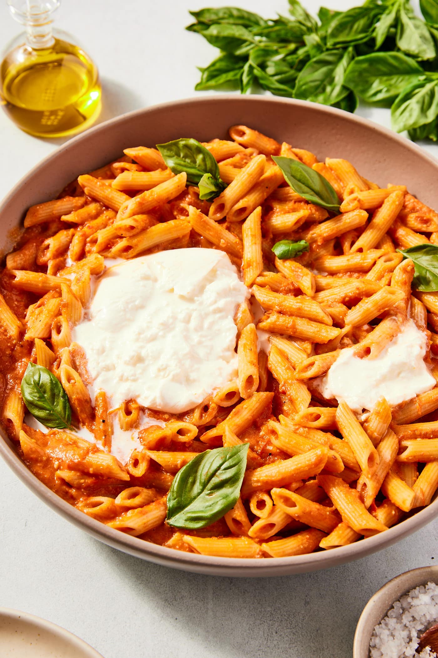 Large brown stoneware bowl filled with roasted tomato pasta sitting on gray stone surface with ball of torn burrata in the middle along with basil leaves.