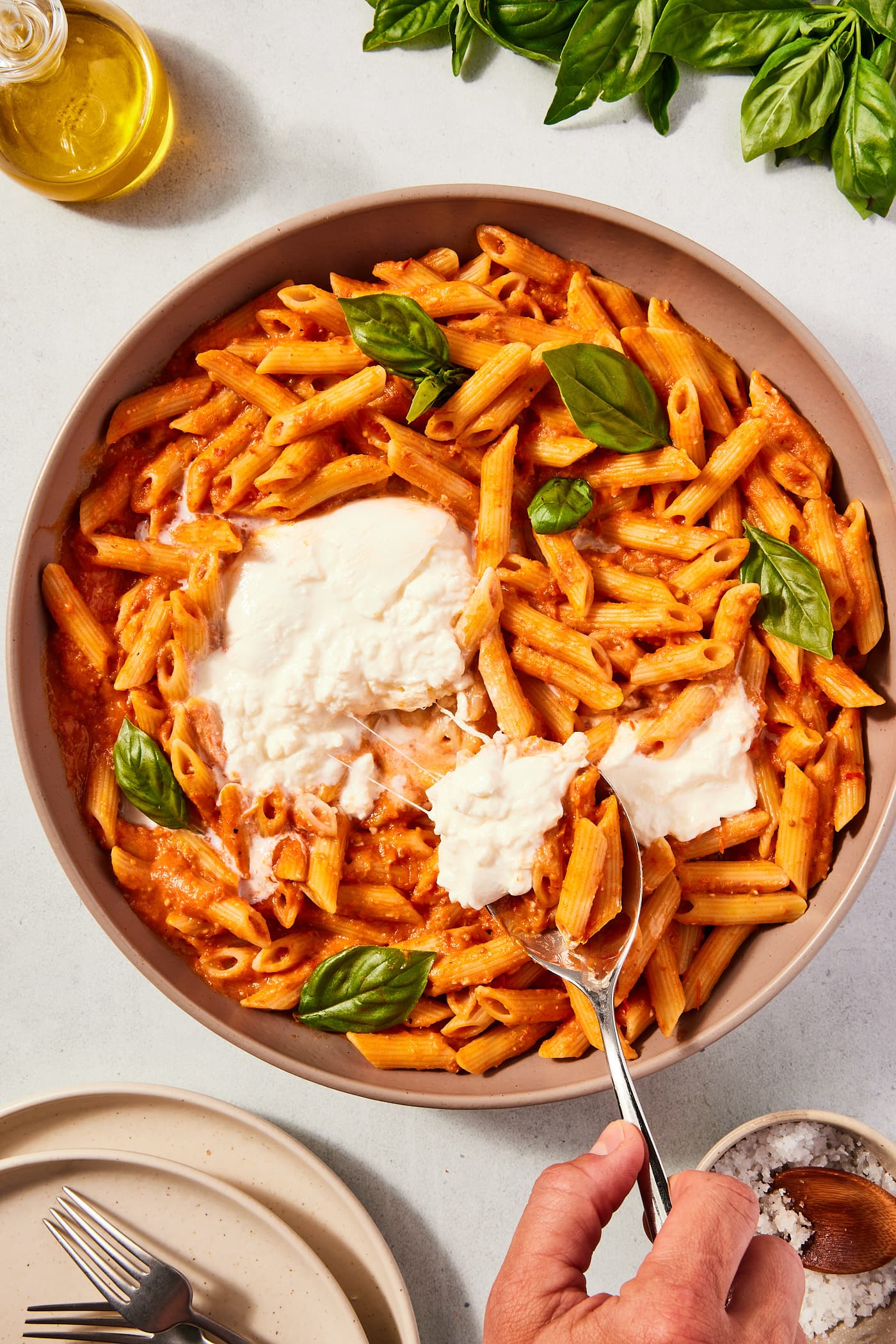Large stoneware bowl filled with pasta covered in roasted tomato sauce with hand holding spoon pulling serving out of the bowl with burrata and cheese strings hanging.
