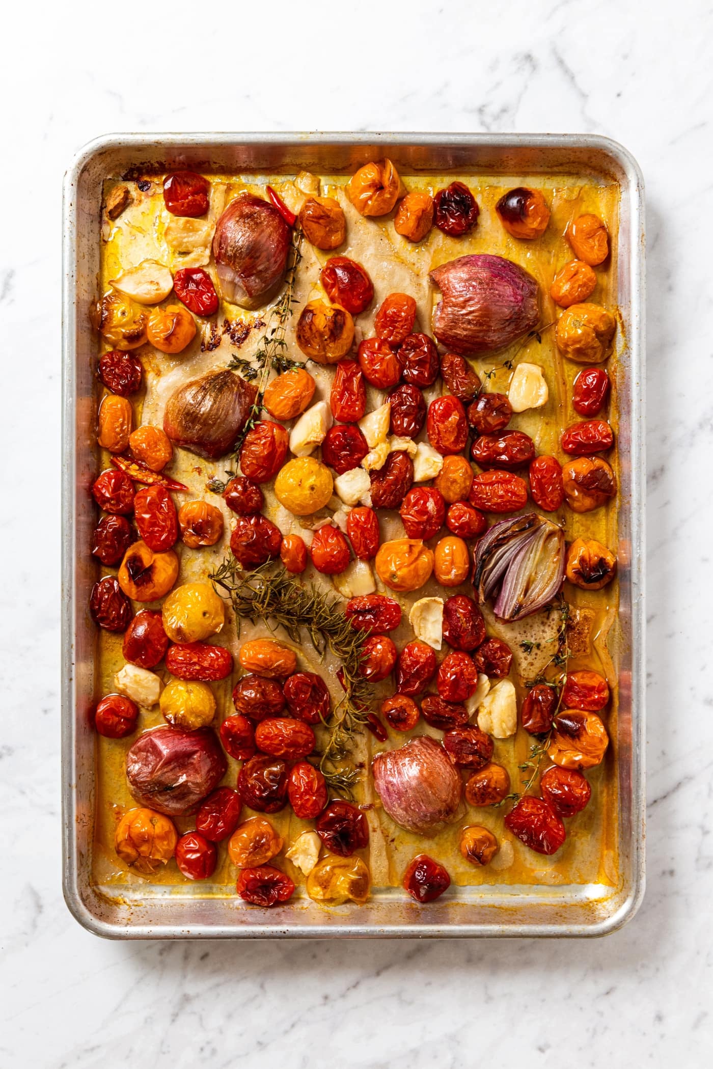 White marble surface with parchment-lined baking sheet filled with roasted cherry tomatoes, shallots, garlic cloves, and herbs right after being removed from the oven.