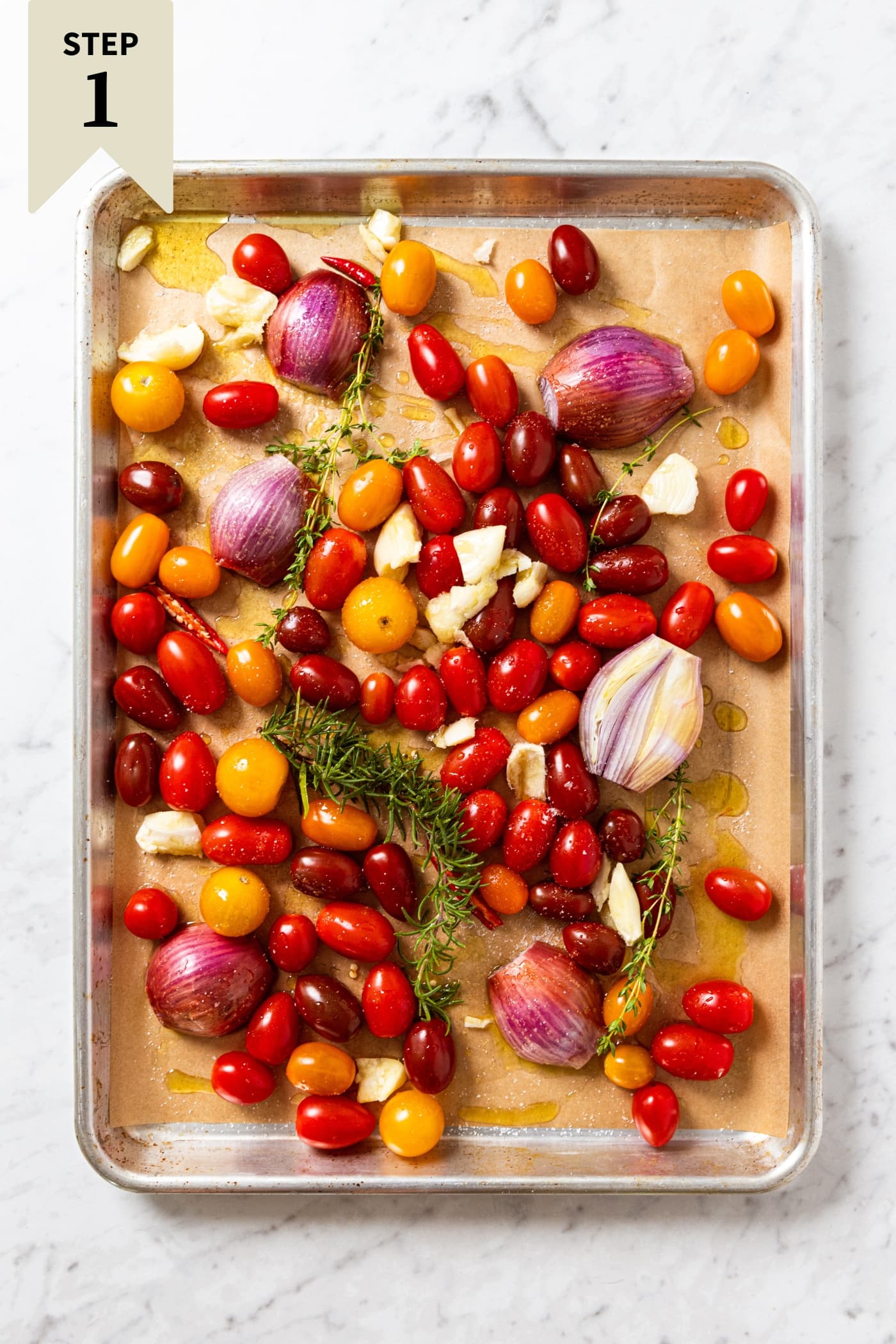 White marble surface with sheet pan filled with raw cherry tomatoes, shallots, and herbs all on piece of parchment right before going into the oven.