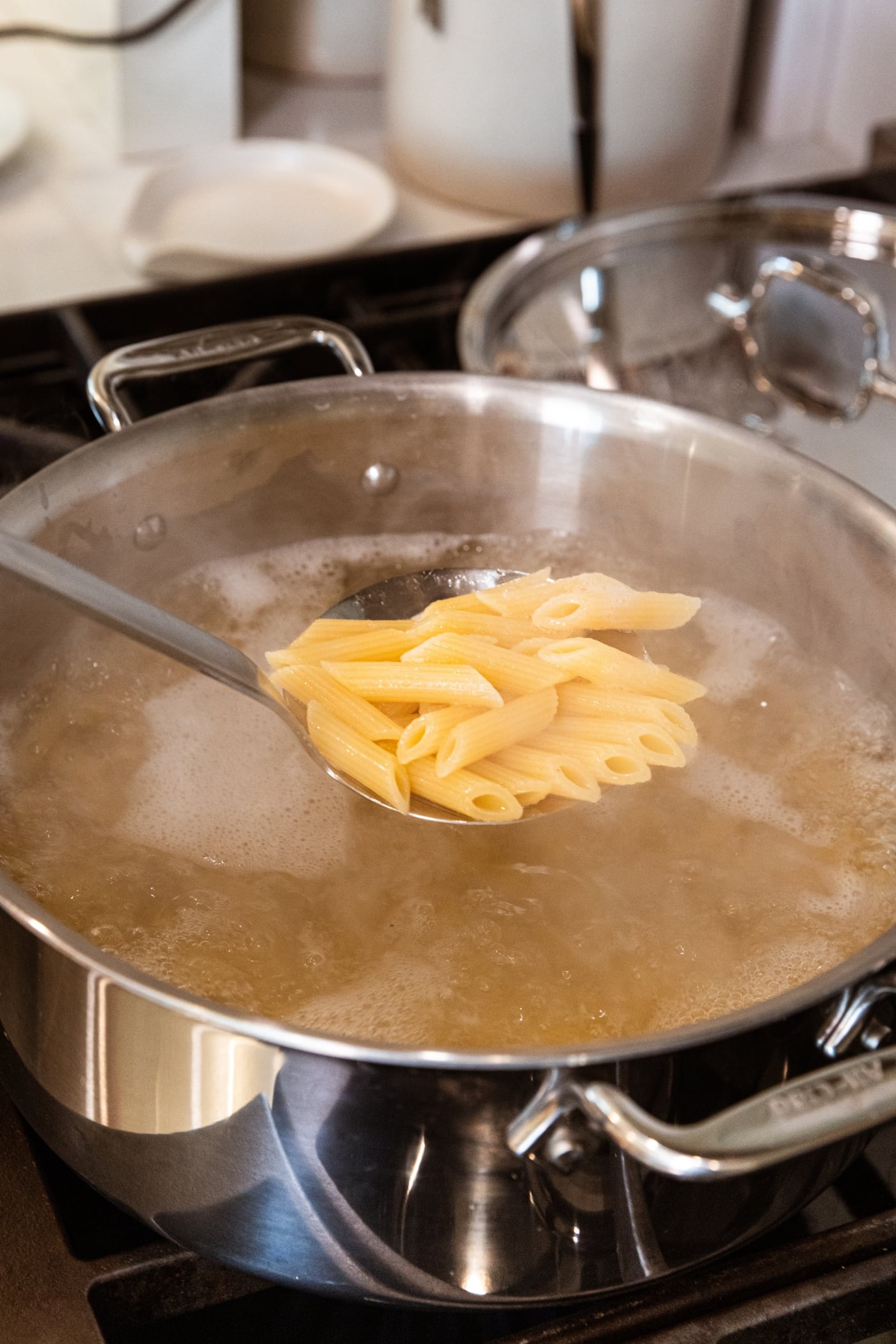 Hand holding metal spoon lifting cooked pasta from a large kettle with boiling water set on the stovetop.