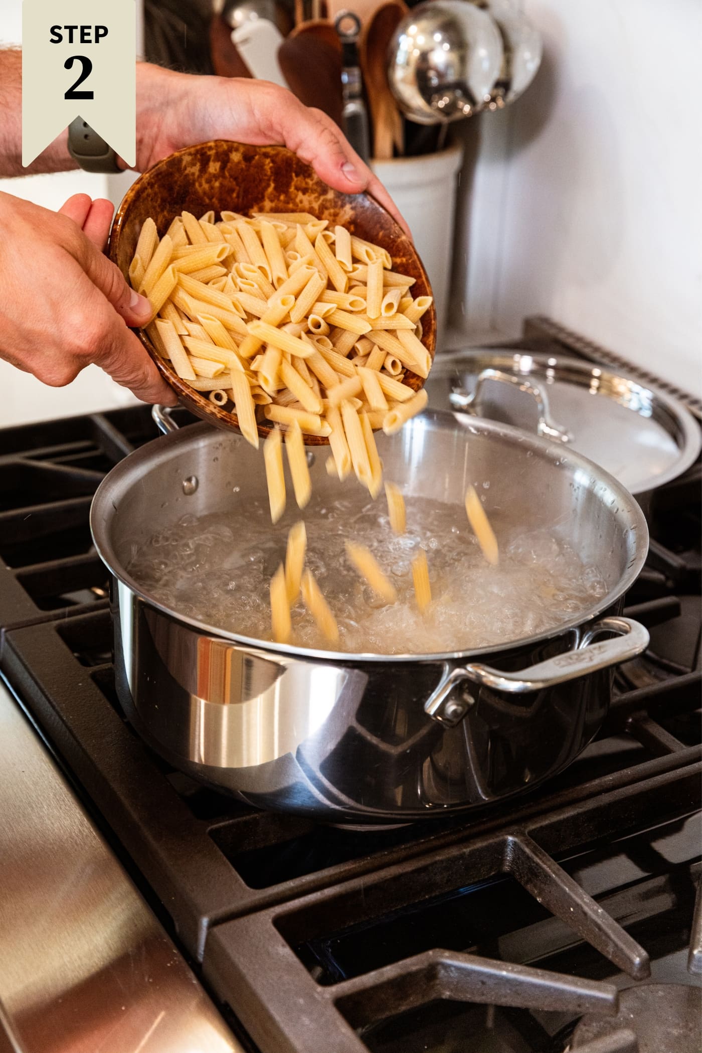 Two hands holding brown bowl, pouring uncooked pasta into boiling water in kettle on stovetop.