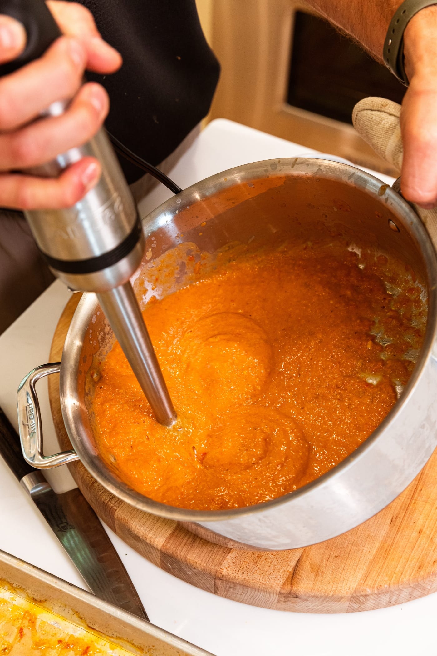 Hand holding handheld immersion blender with blended orange-colored tomato sauce made from roasted tomatoes in large kettle.