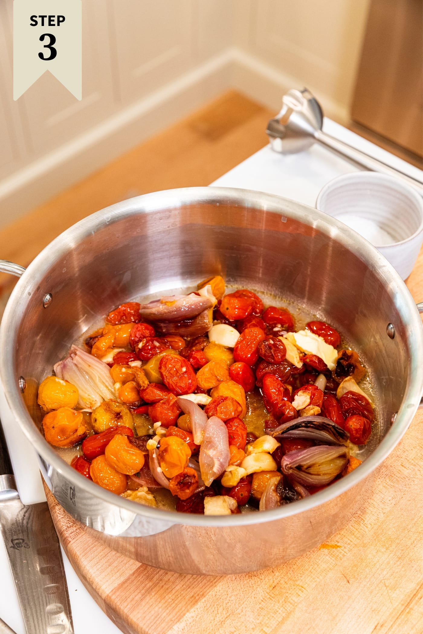 Large kettle sitting on wood cutting board filled with roasted tomatoes right before being blended together with handheld immersion blender.