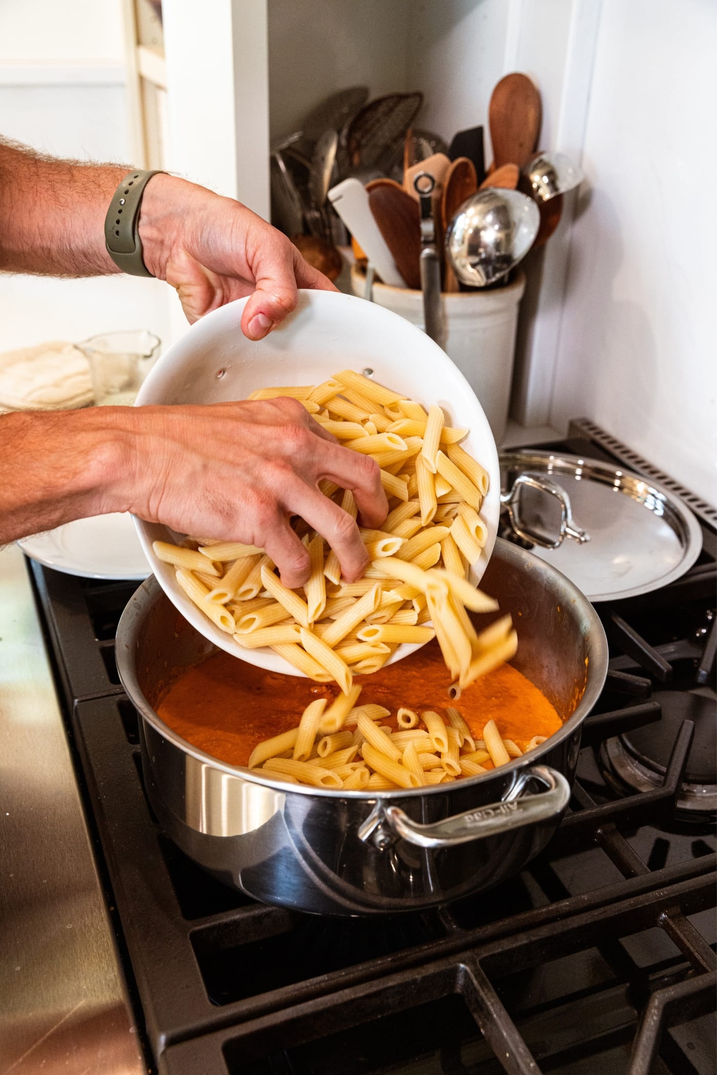 Hand pouring cooked penne pasta into large kettle filled with roasted tomato sauce on the stovetop.