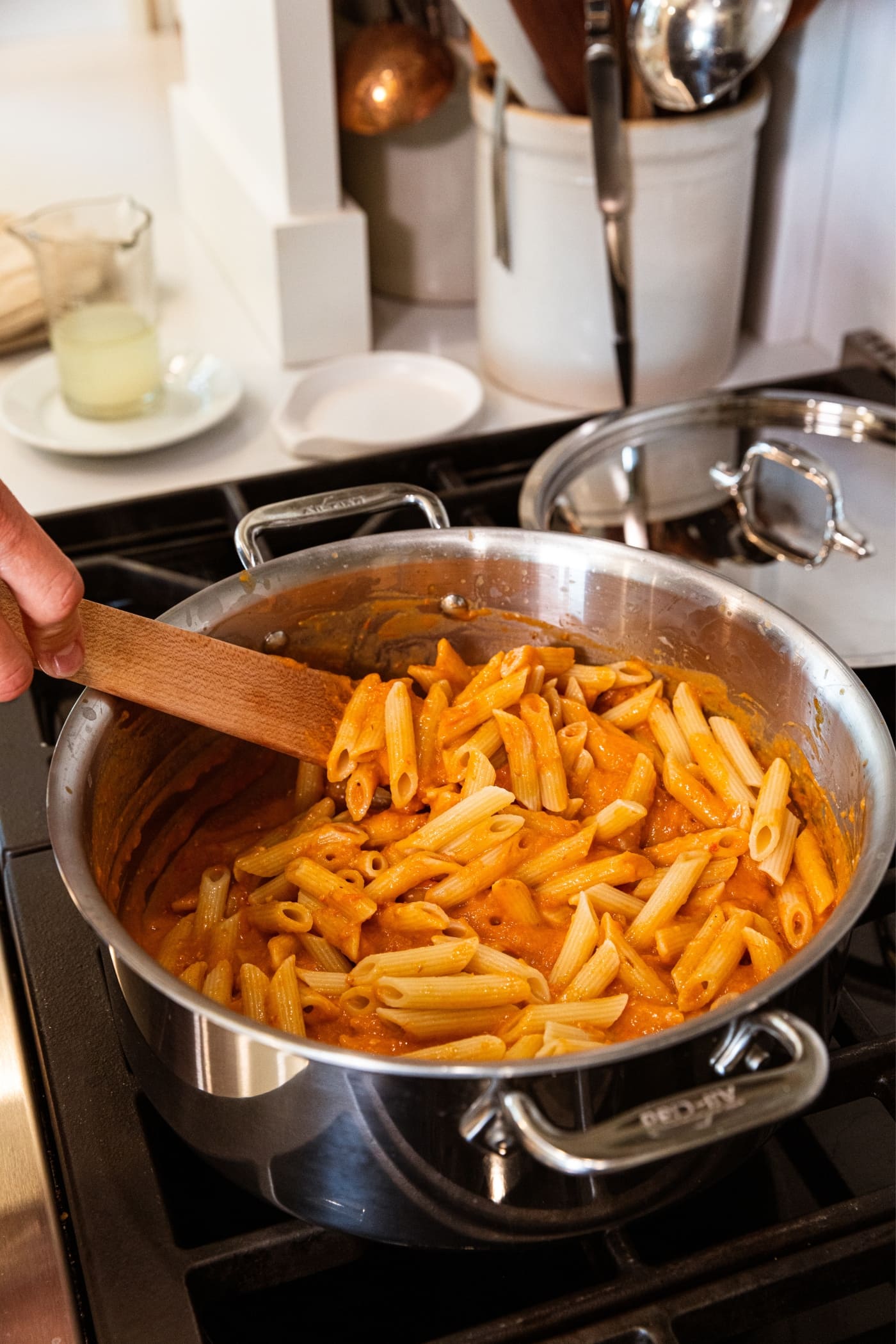 Hand holding wooden utensil mixing together cooked penne pasta with roasted tomato sauce in large kettle on the stovetop.