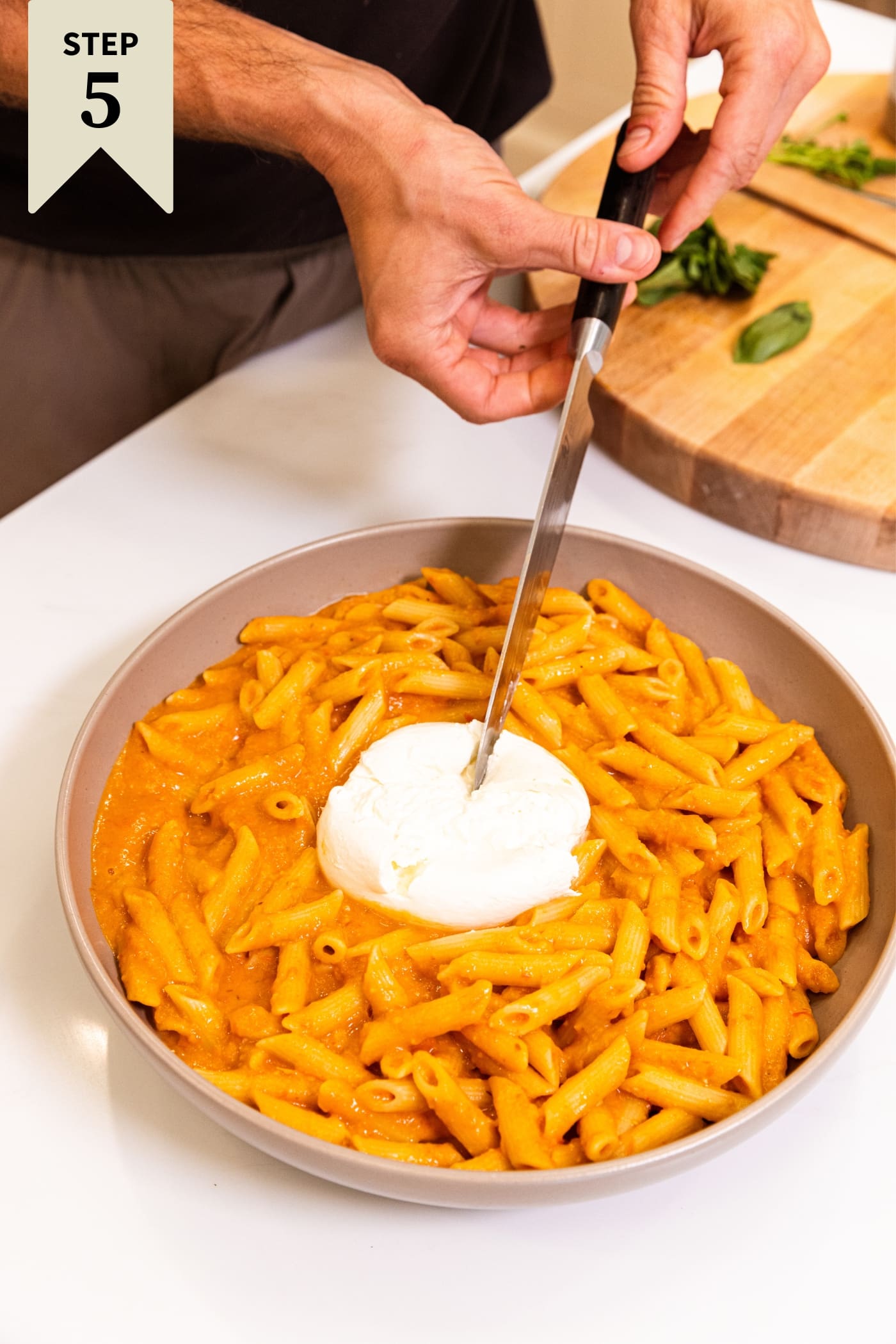 Hand holding knife slicing into ball of burrata in the middle of a large bowl of pasta on white surface.