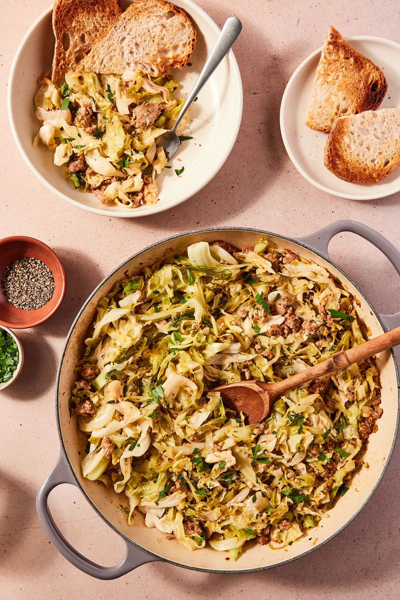 Large skillet filled with cooked cabbage and sausage with wooden spoon scooping up a serving with a serving already in a bowl with toasted slices of bread.