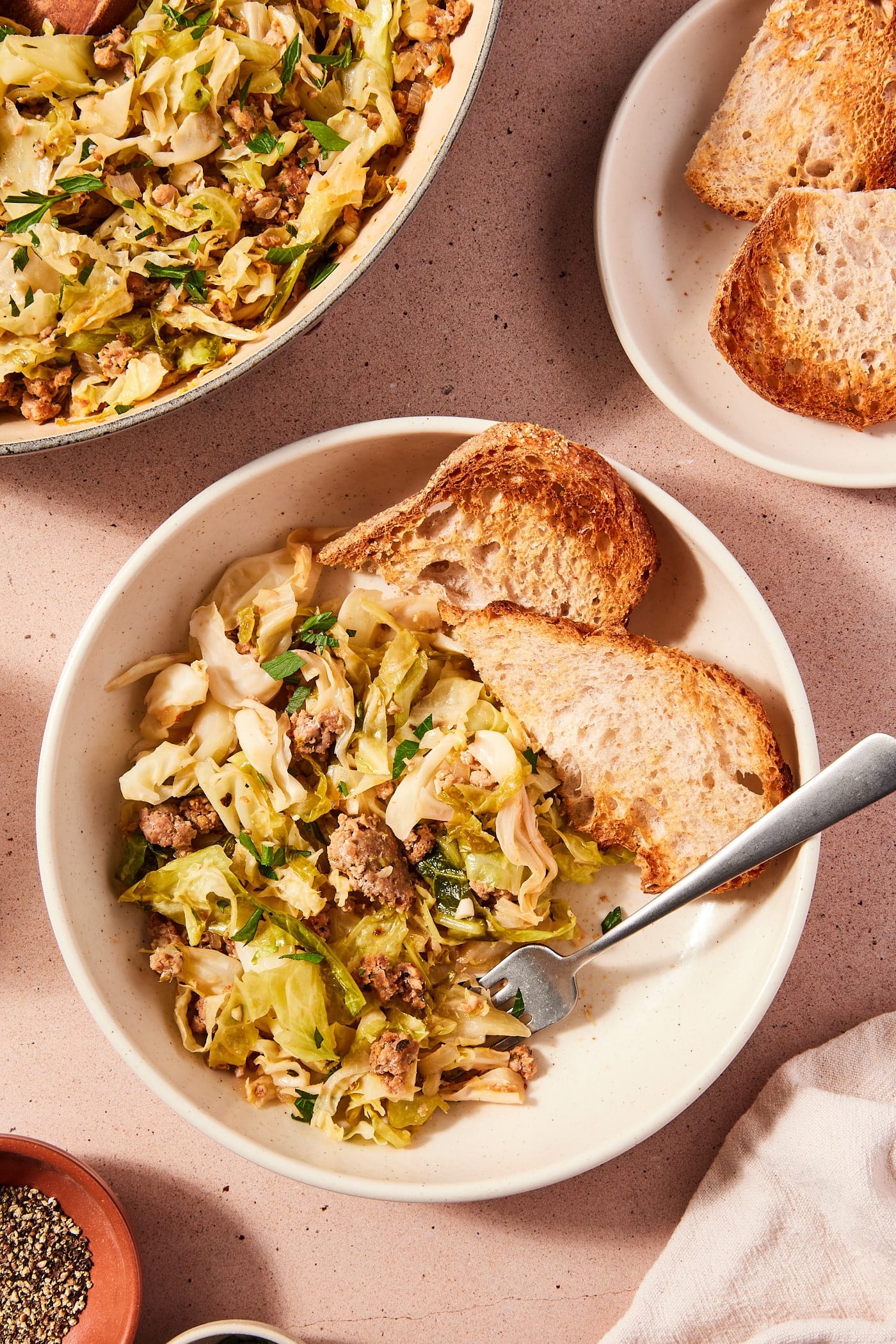 White bowl filled with serving of skillet sausage and cabbage with slices of toasted bread alongside with skillet filled with rest of the cooked cabbage.